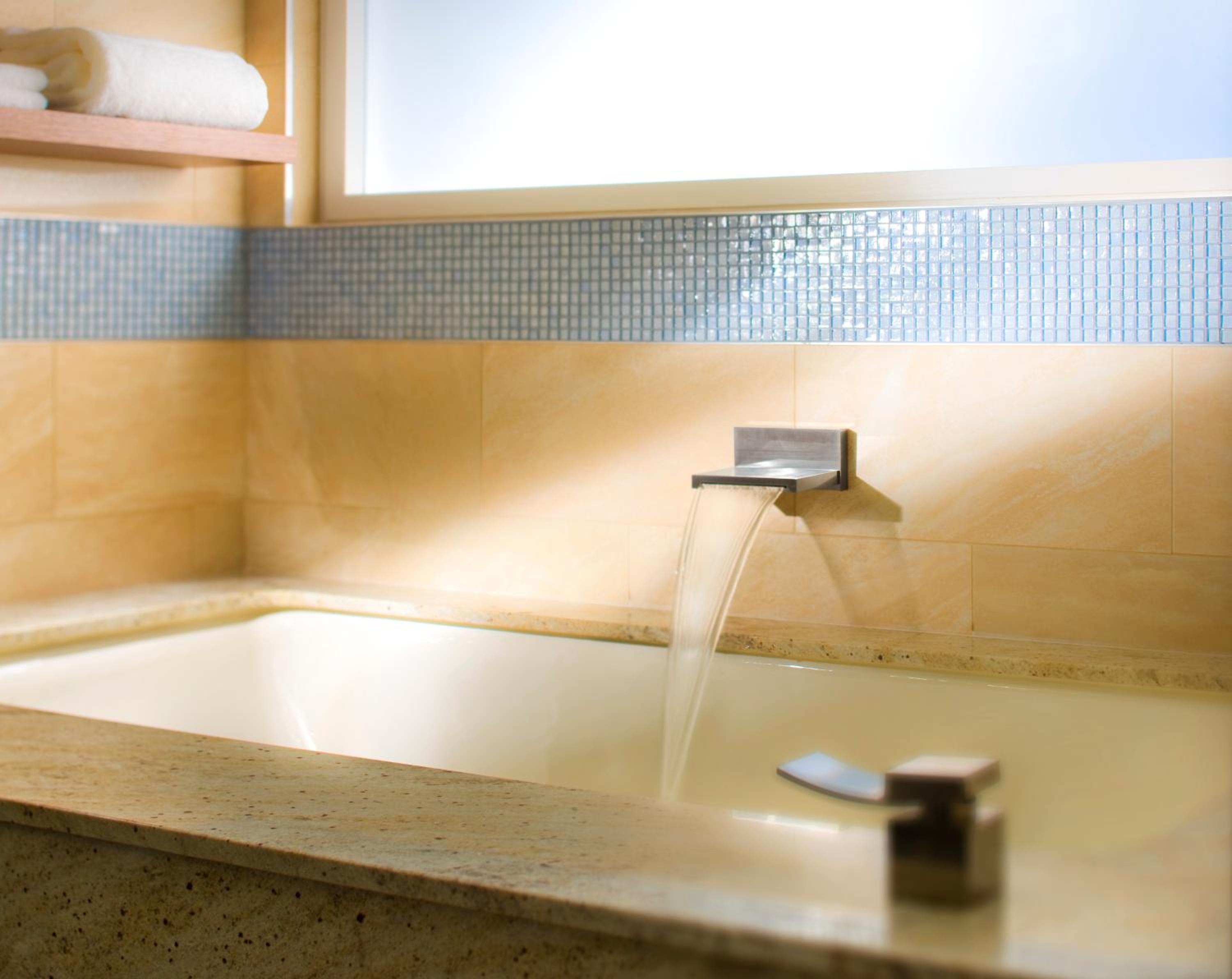 Water cascades from a waterfall spout into a built-in bathtub surrounded by tan stone and blue mosaic tiles.
