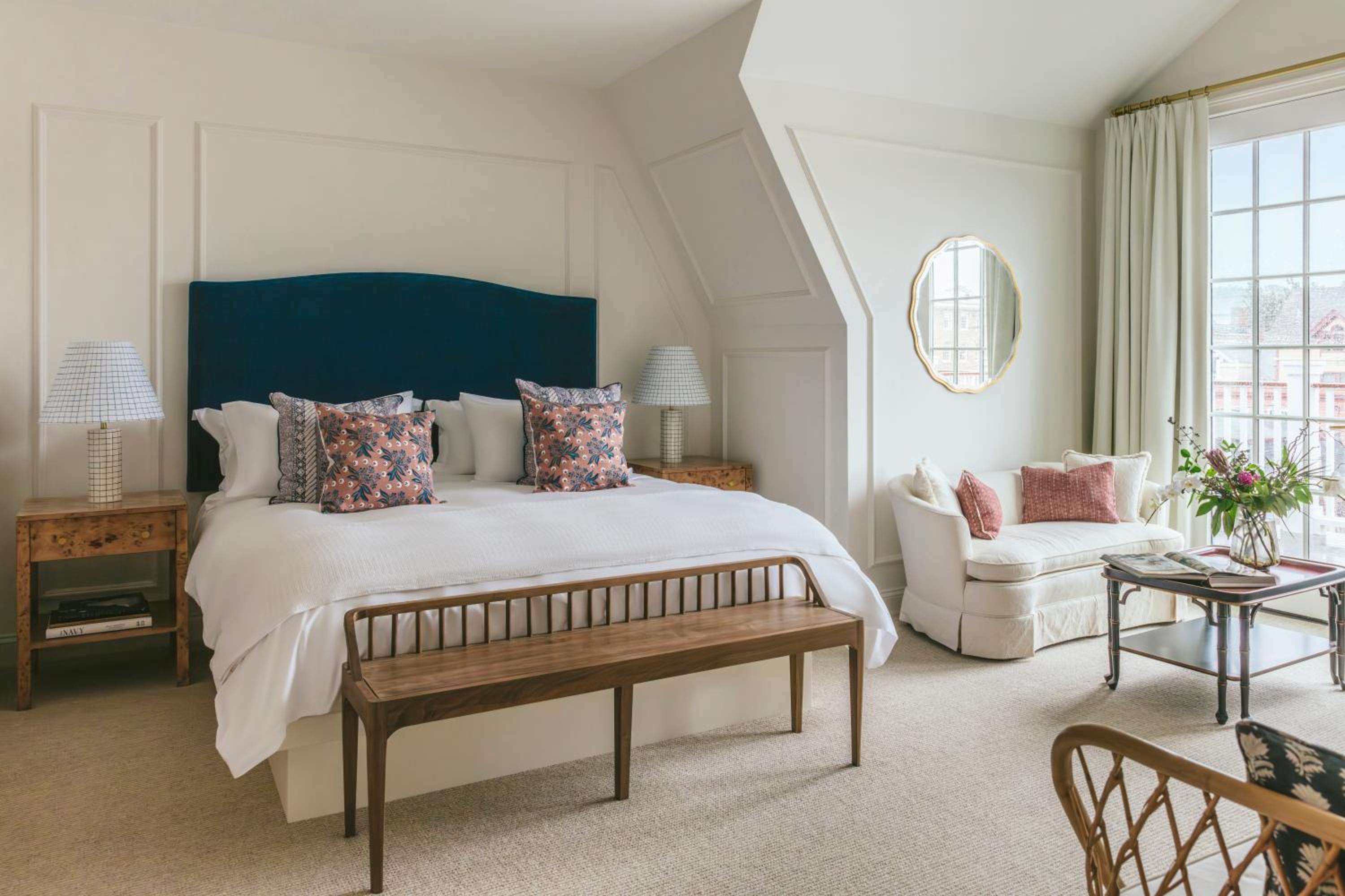 A bedroom with a dark teal headboard, patterned pillows, a wooden bench, and a small sofa beside French doors.