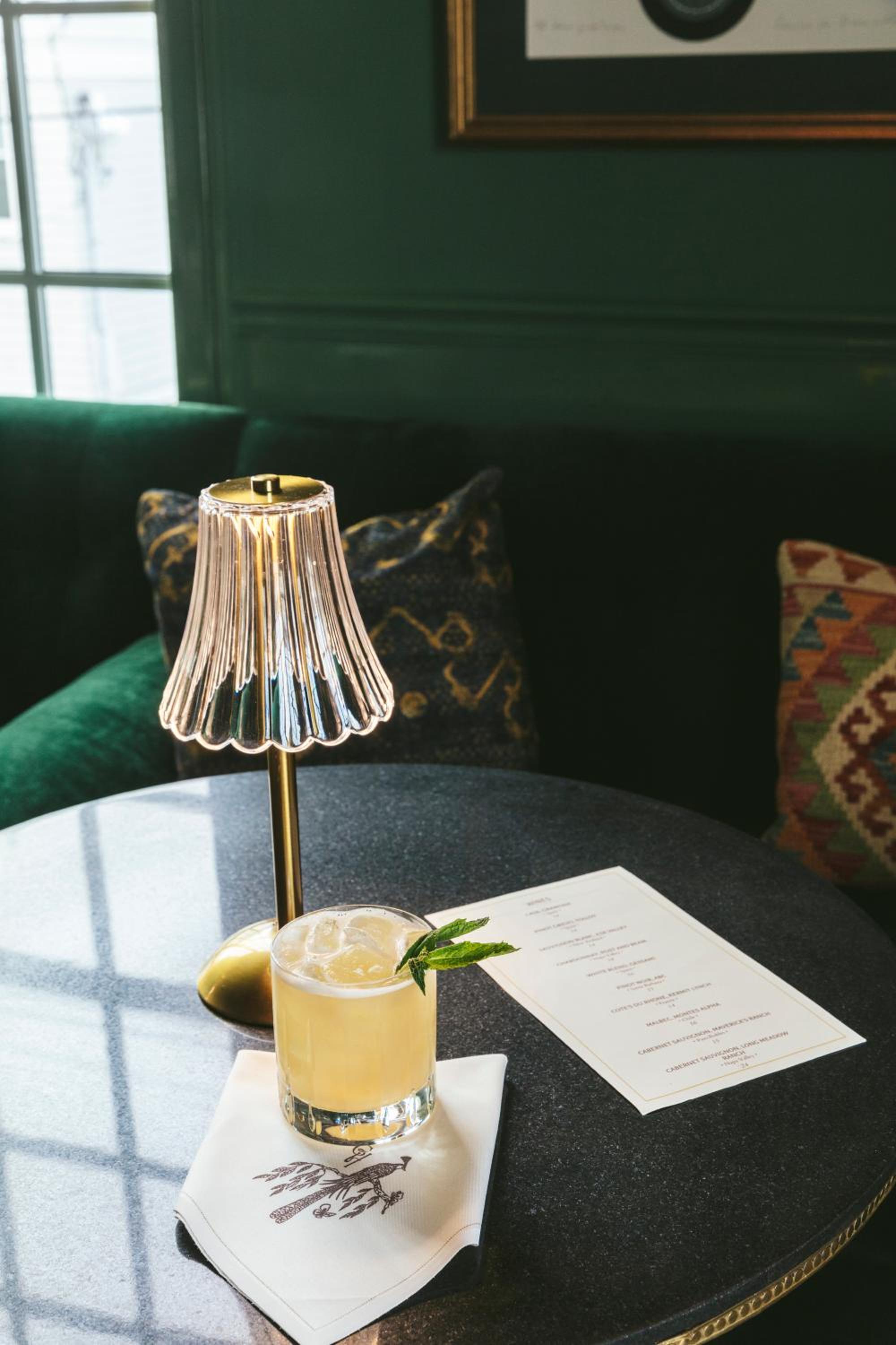 An iced cocktail garnished with mint beside a glowing ribbed table lamp on a dark round tabletop.