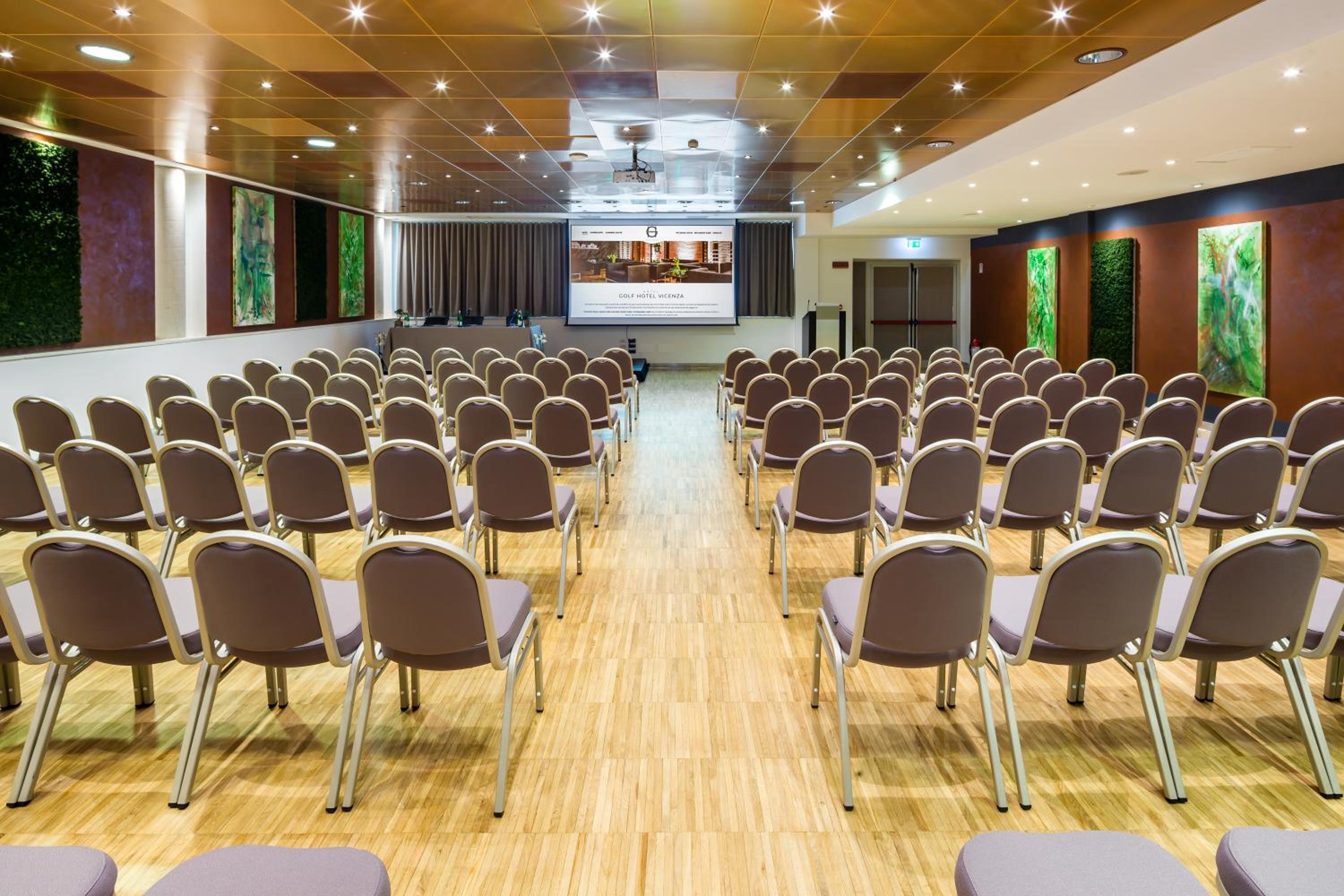 Conference hall with rows of upholstered chairs facing a projection screen over parquet flooring.