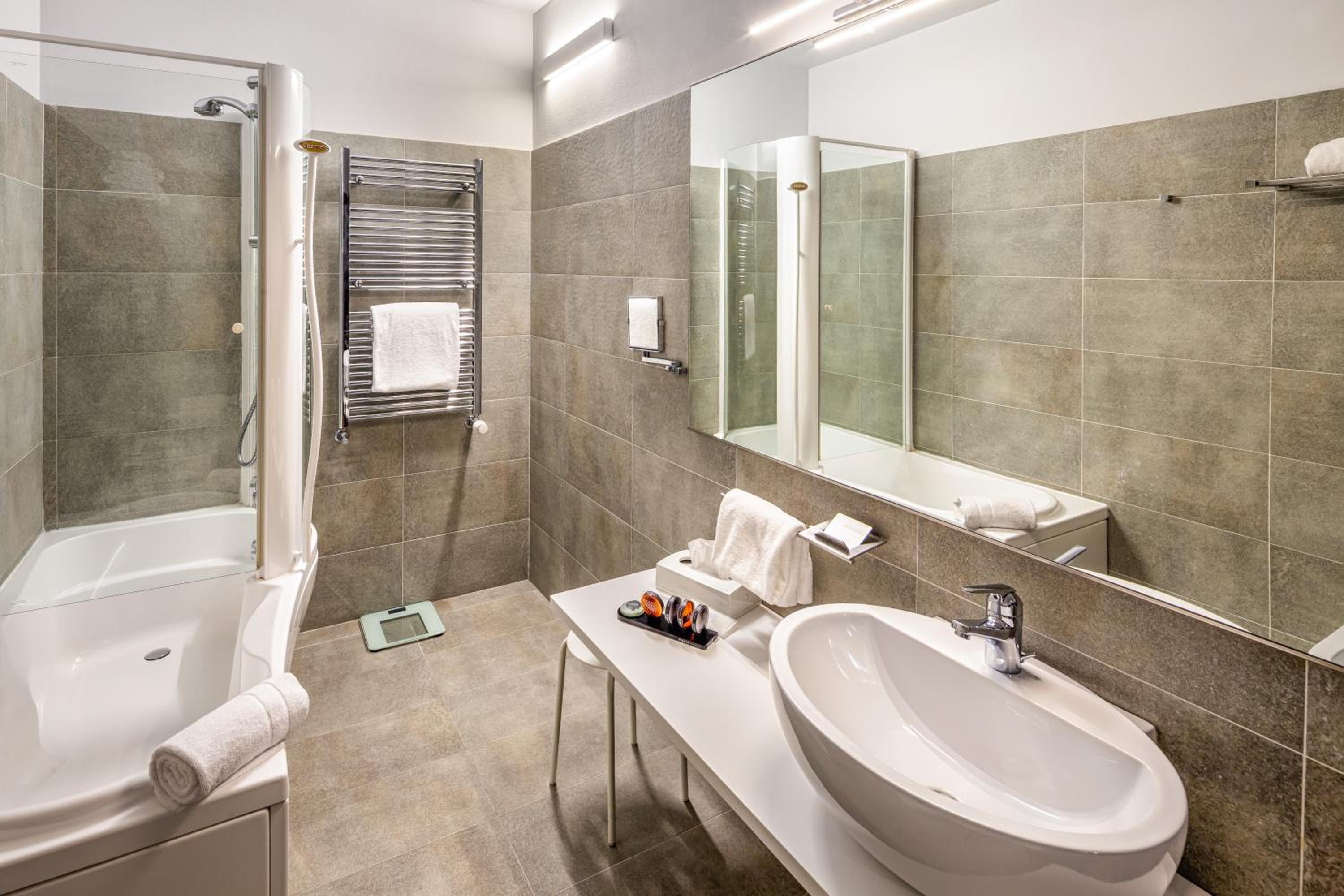 Bathroom with grey tile walls, a wide mirror, oval sink, heated towel rail, and combination bathtub shower.