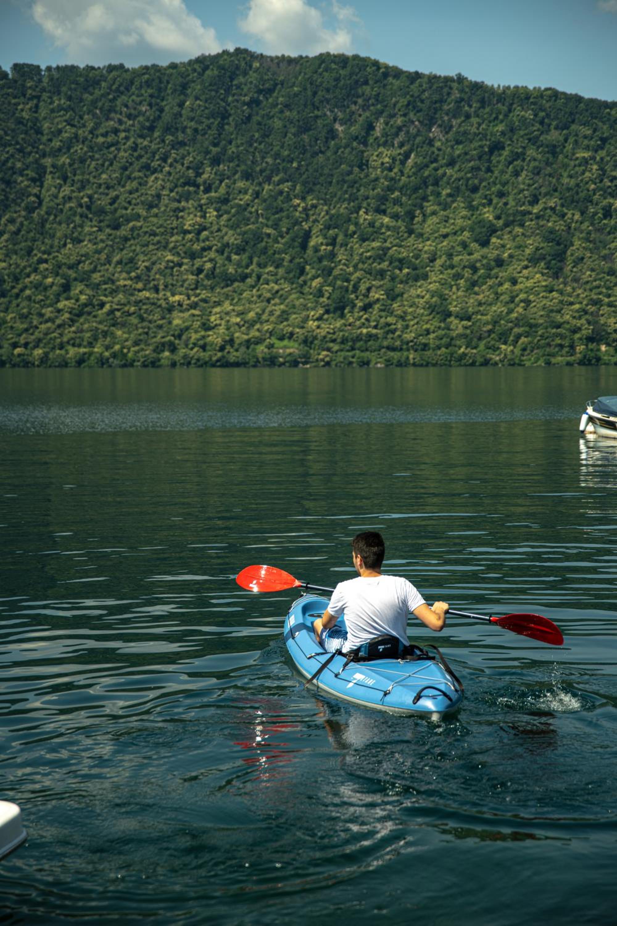 A kayaker paddling across calm lake water toward a steep, tree-covered mountain.