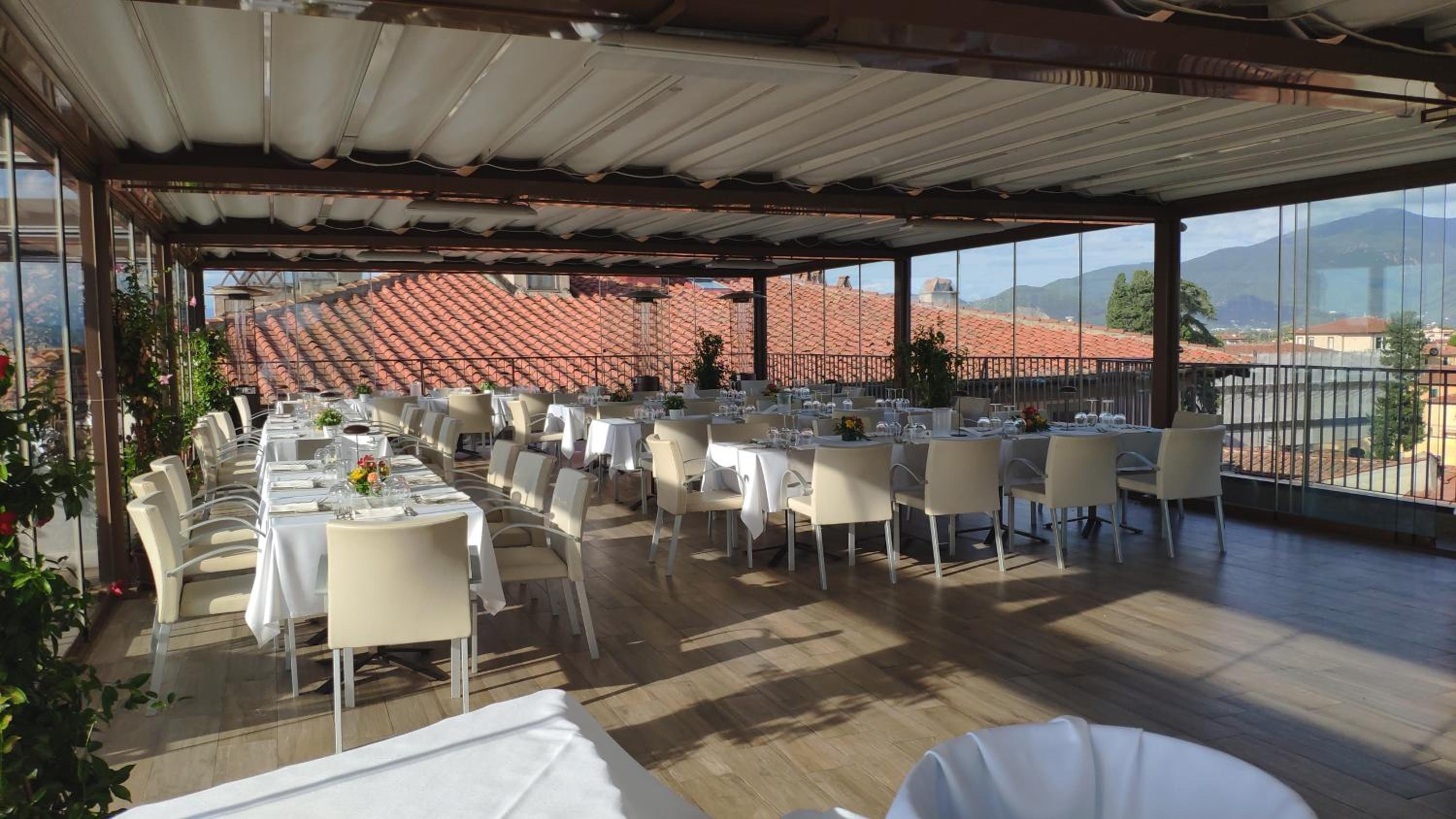 A covered rooftop terrace with dining tables, wood-look floors, and terracotta rooftops through glass walls.