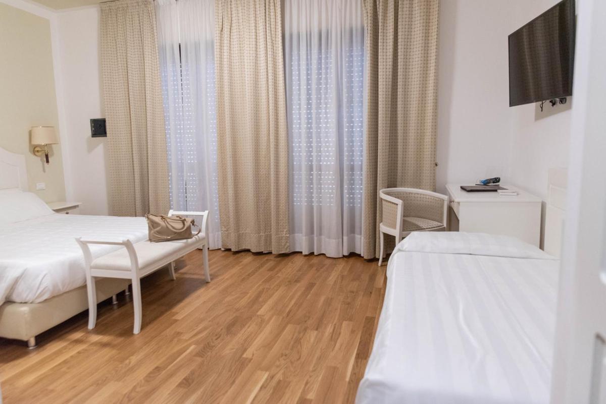 Guest bedroom with wood flooring, white linens, a cushioned bench, and tall window drapes.