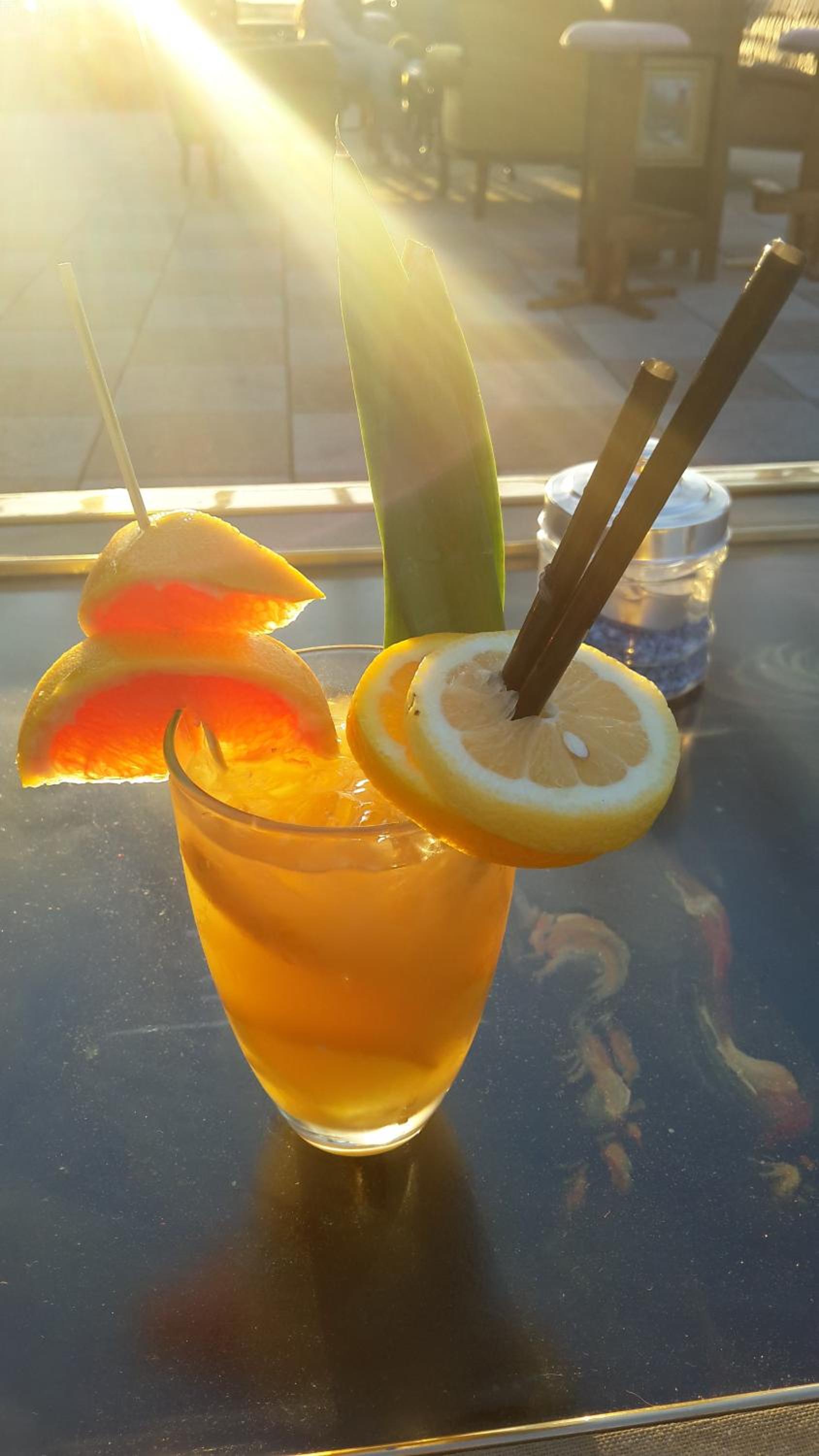 An iced cocktail garnished with citrus slices and straws on an outdoor table in bright sunlight.