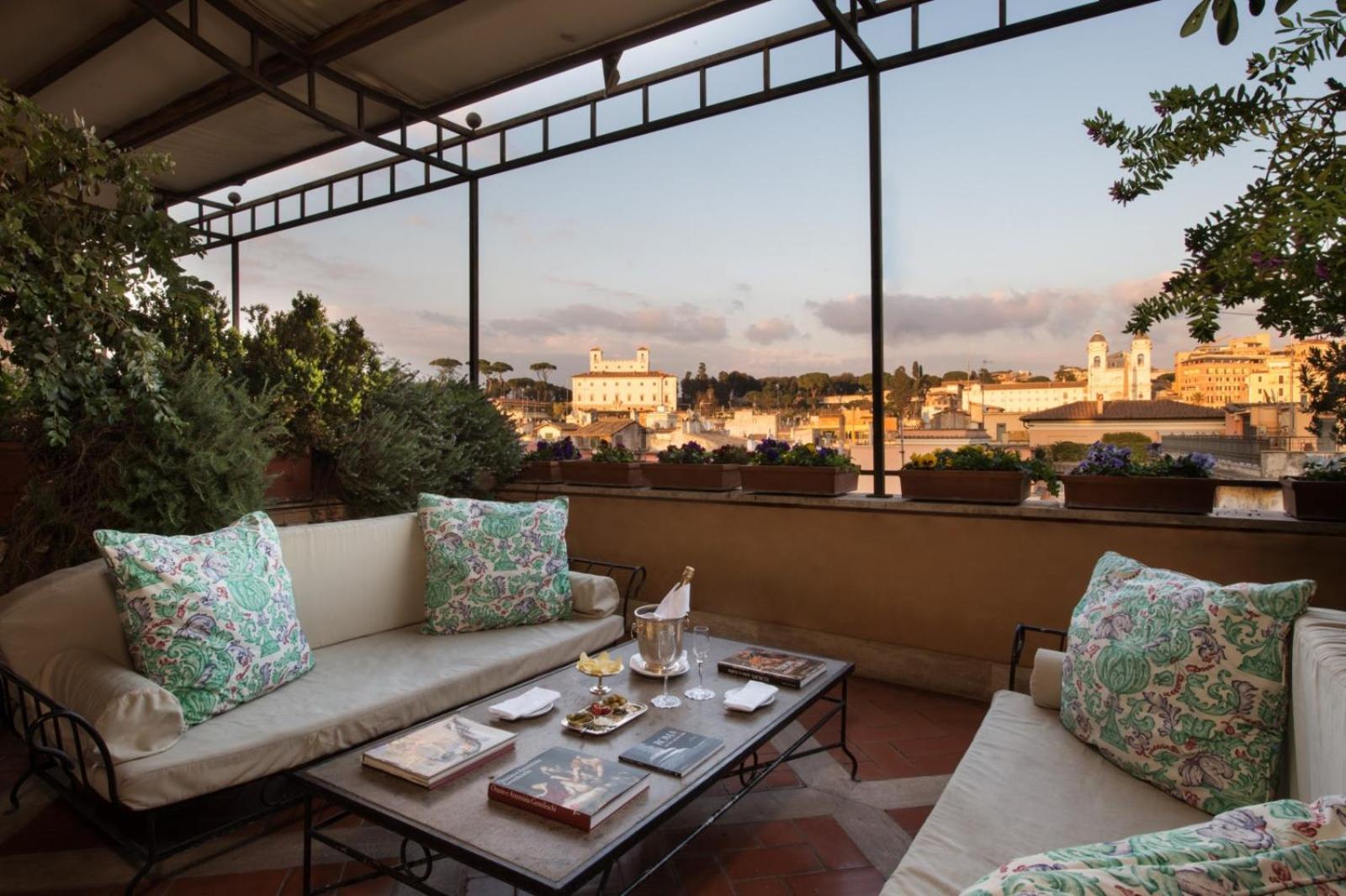 A rooftop terrace with cushioned sofas, a low coffee table, potted flowers, and views across the city skyline.