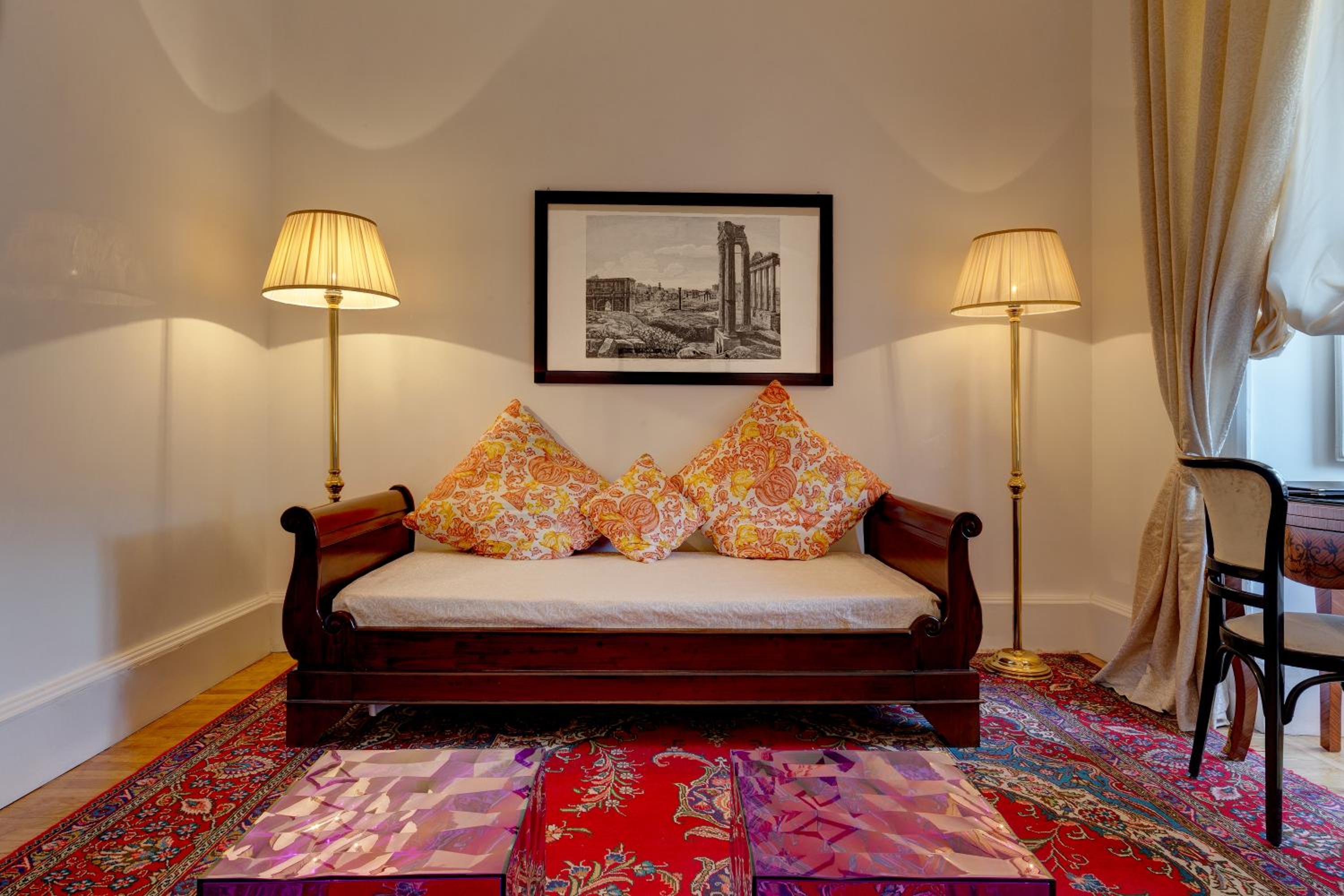 A sitting area with a wooden daybed, patterned pillows, standing lamps, and a framed print of Roman ruins.