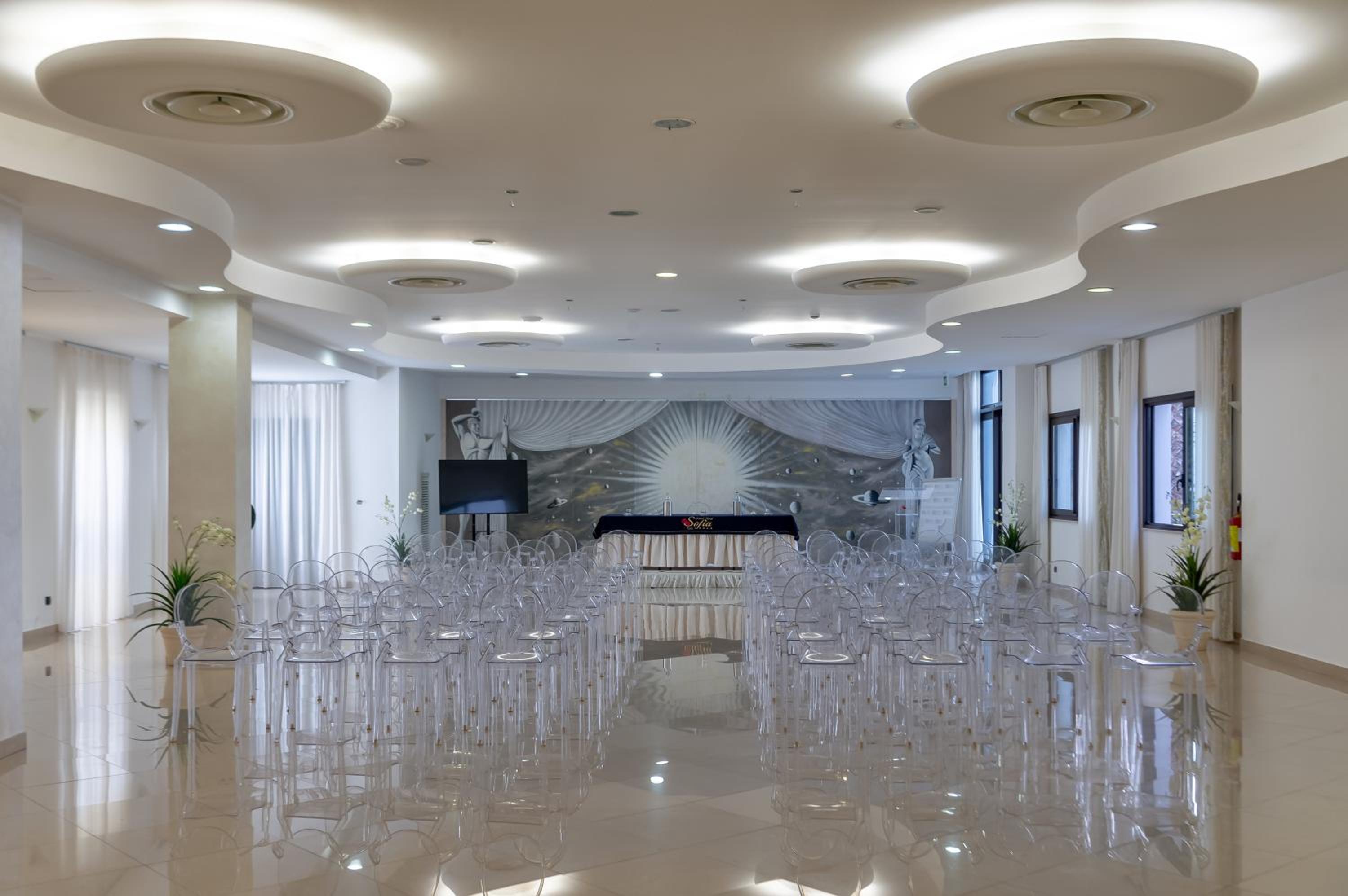 An event hall with clear acrylic chairs facing a presentation table beneath circular recessed ceiling lights.