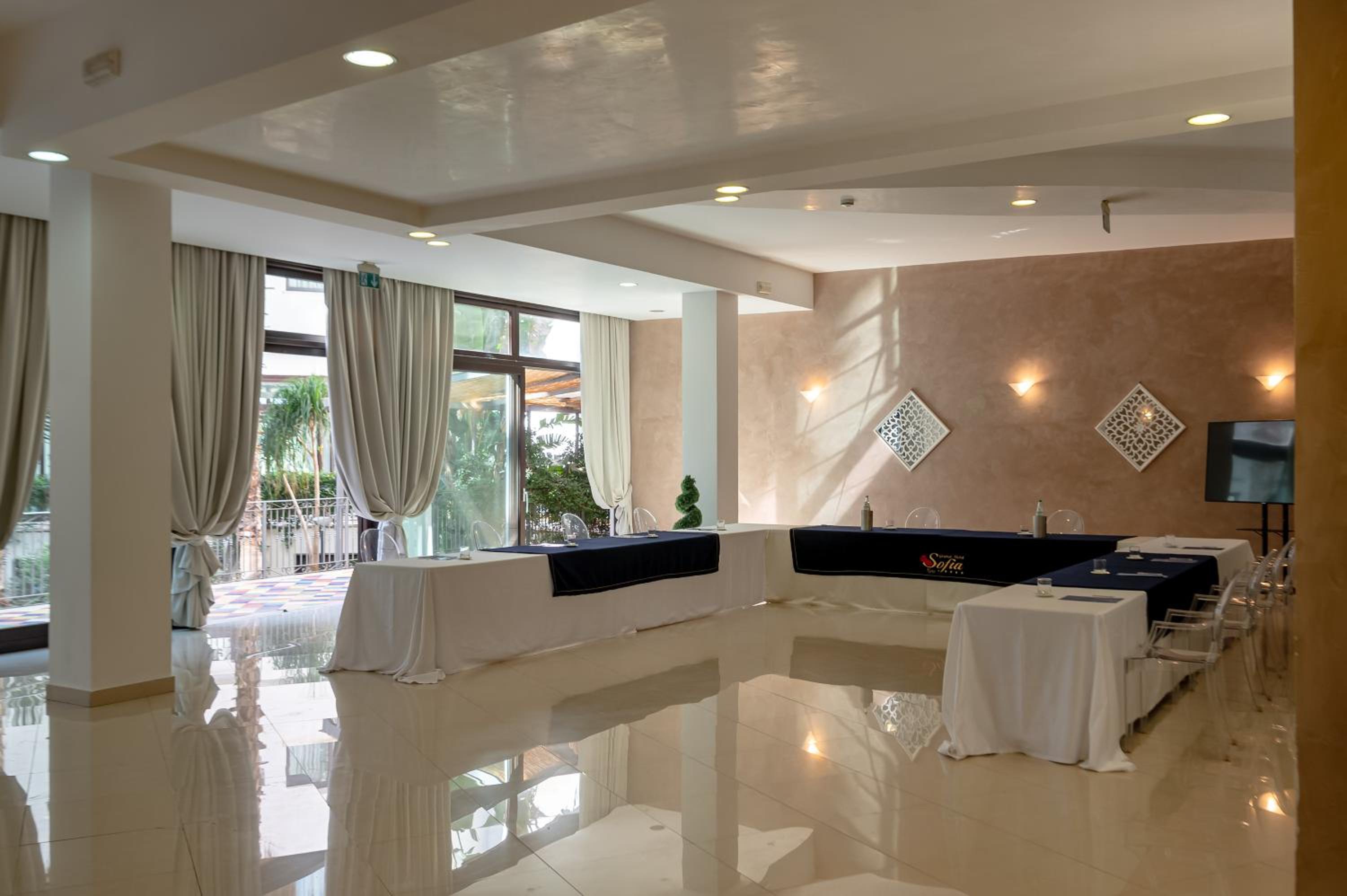 A meeting room with draped tables arranged in a horseshoe shape, clear chairs, and sheer draped windows.