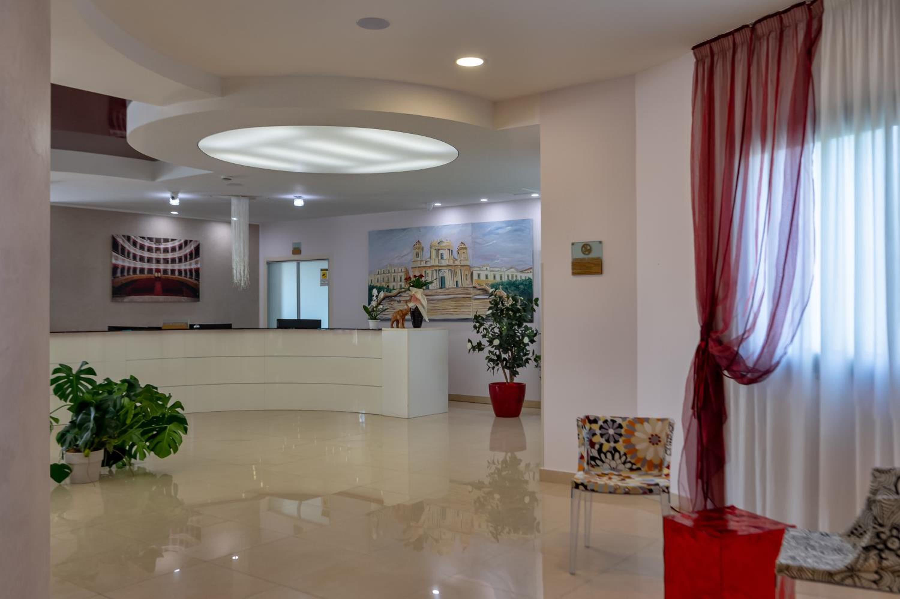 A hotel reception area with a curved white desk, polished floors, potted plants, and sheer red curtains.
