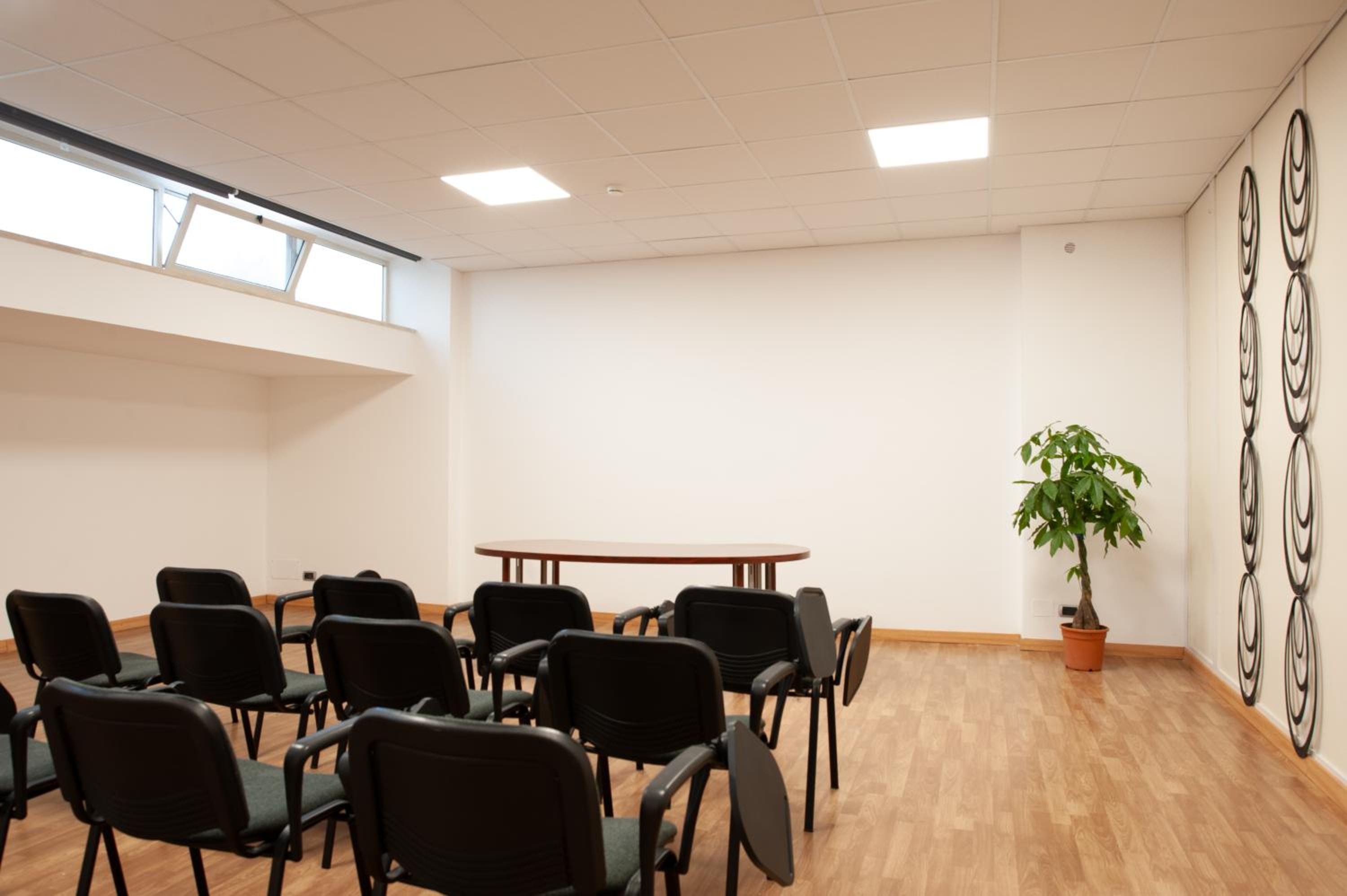 A meeting room with rows of black chairs facing a wooden table, light wood flooring, and a potted plant.