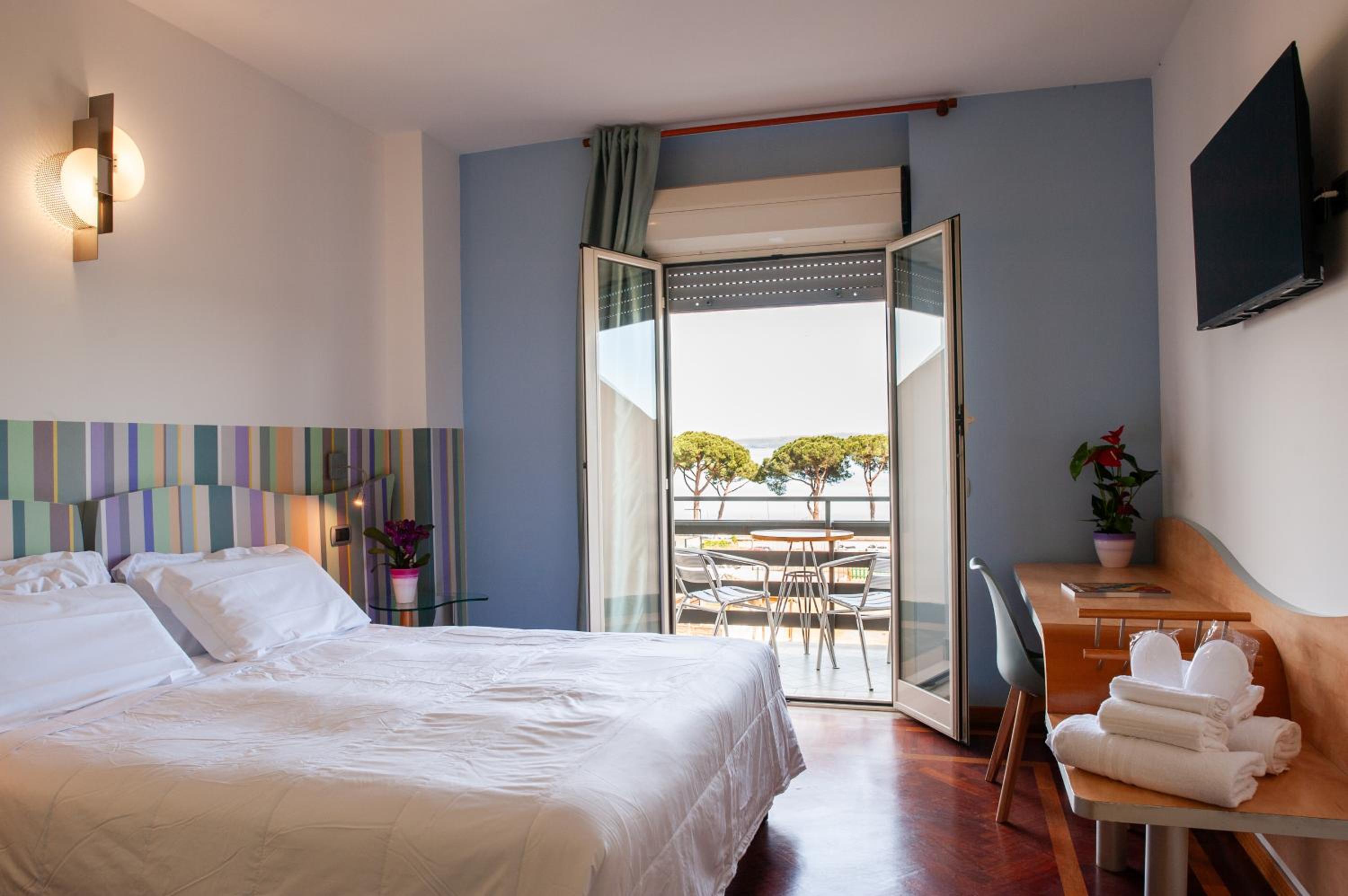 A bedroom with white bedding, wood floors, and open doors leading to a balcony overlooking pine trees and water.