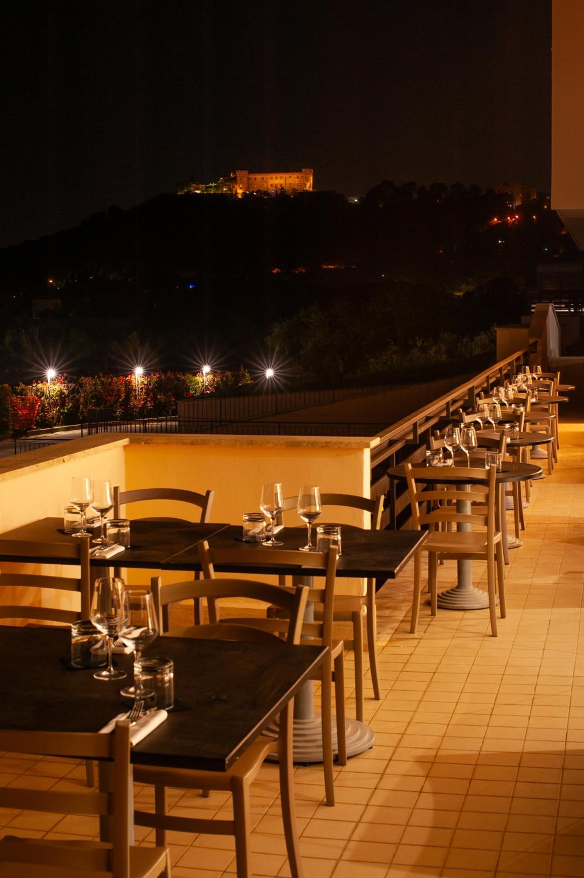 An outdoor terrace at night with dining tables set with glassware, overlooking a lit hilltop fortress.