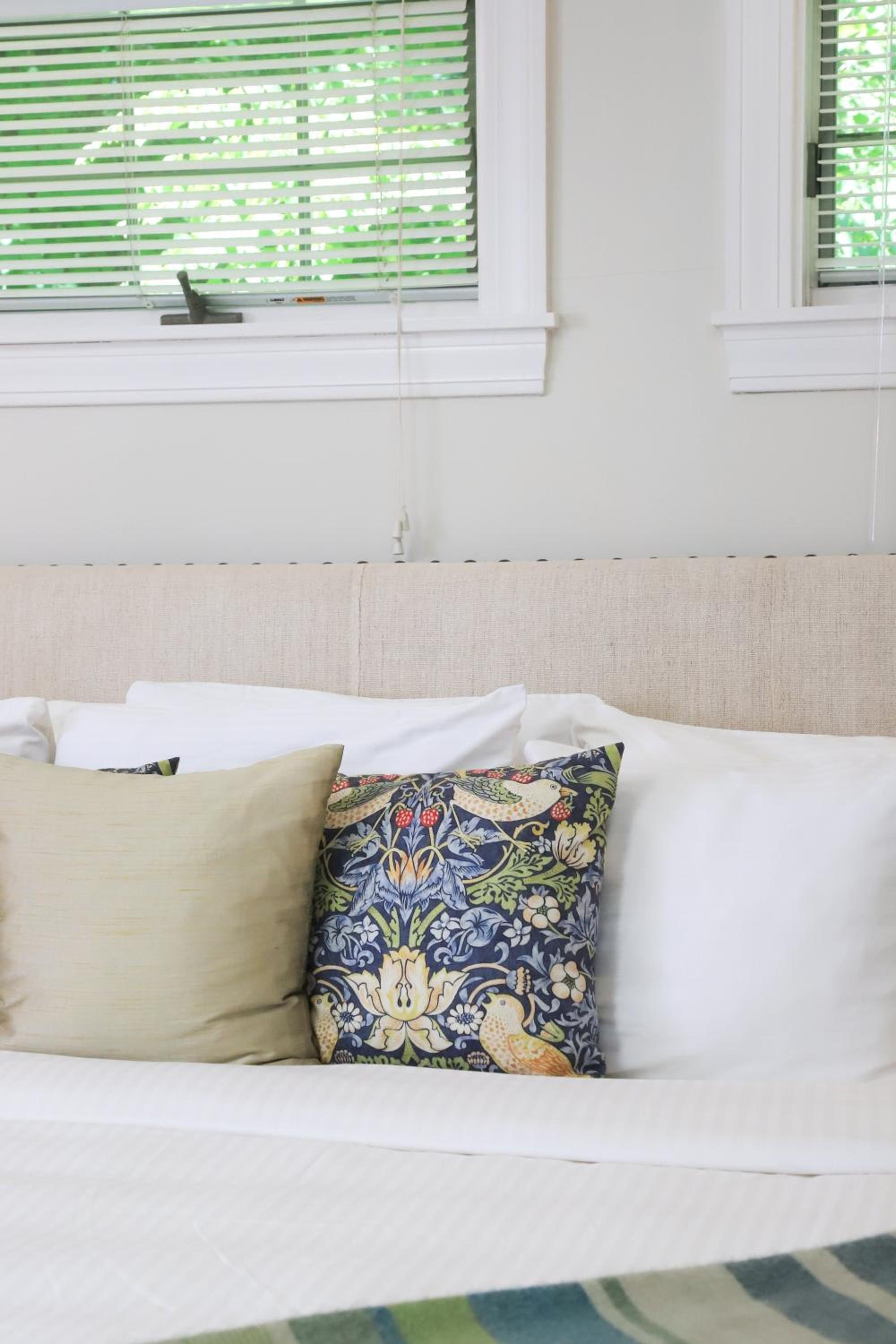 A bed with an upholstered beige headboard, white linens, and a botanical patterned accent pillow below blinds.