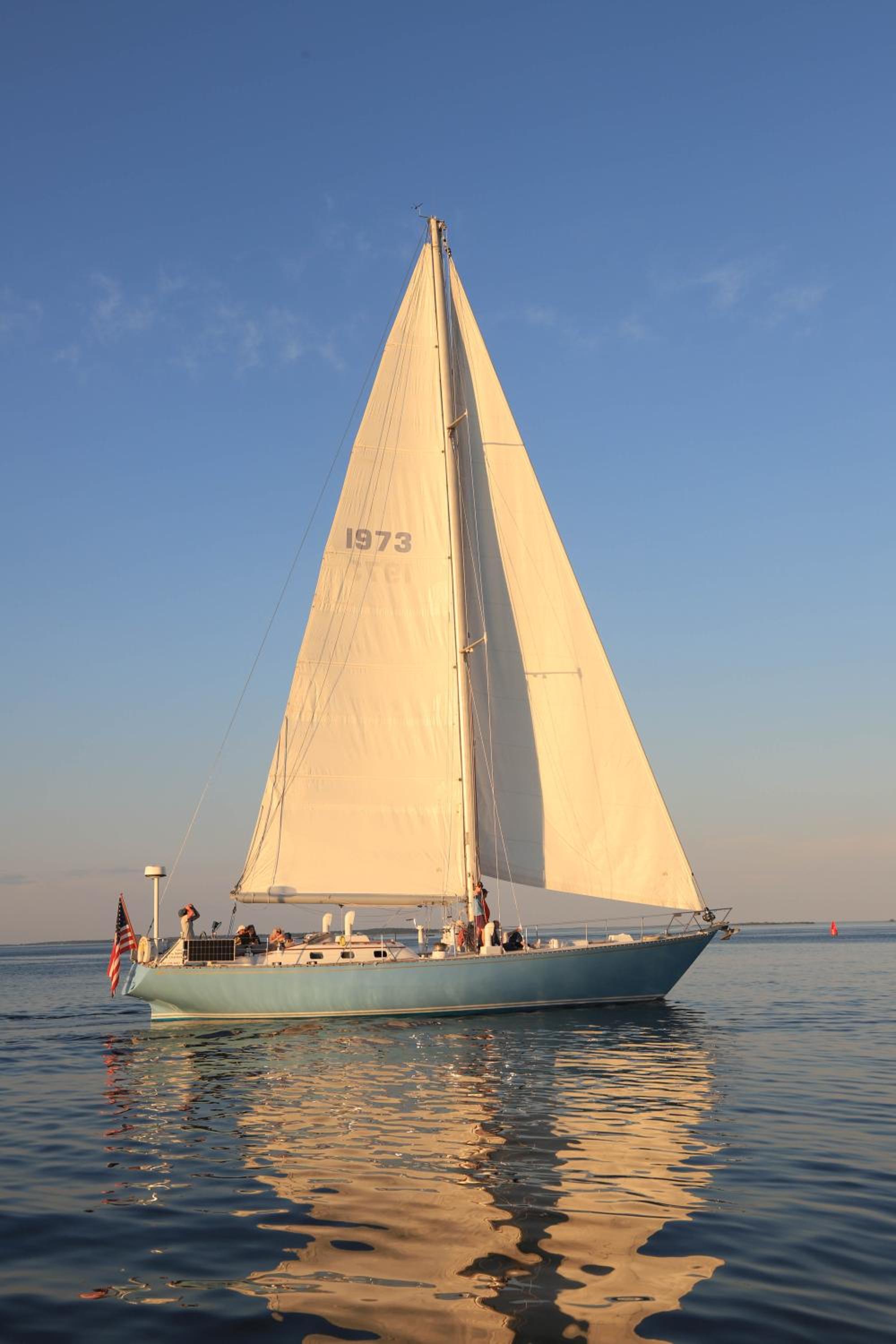 A blue-hulled sailboat with white sails glides across calm sea water under a sunny sky.