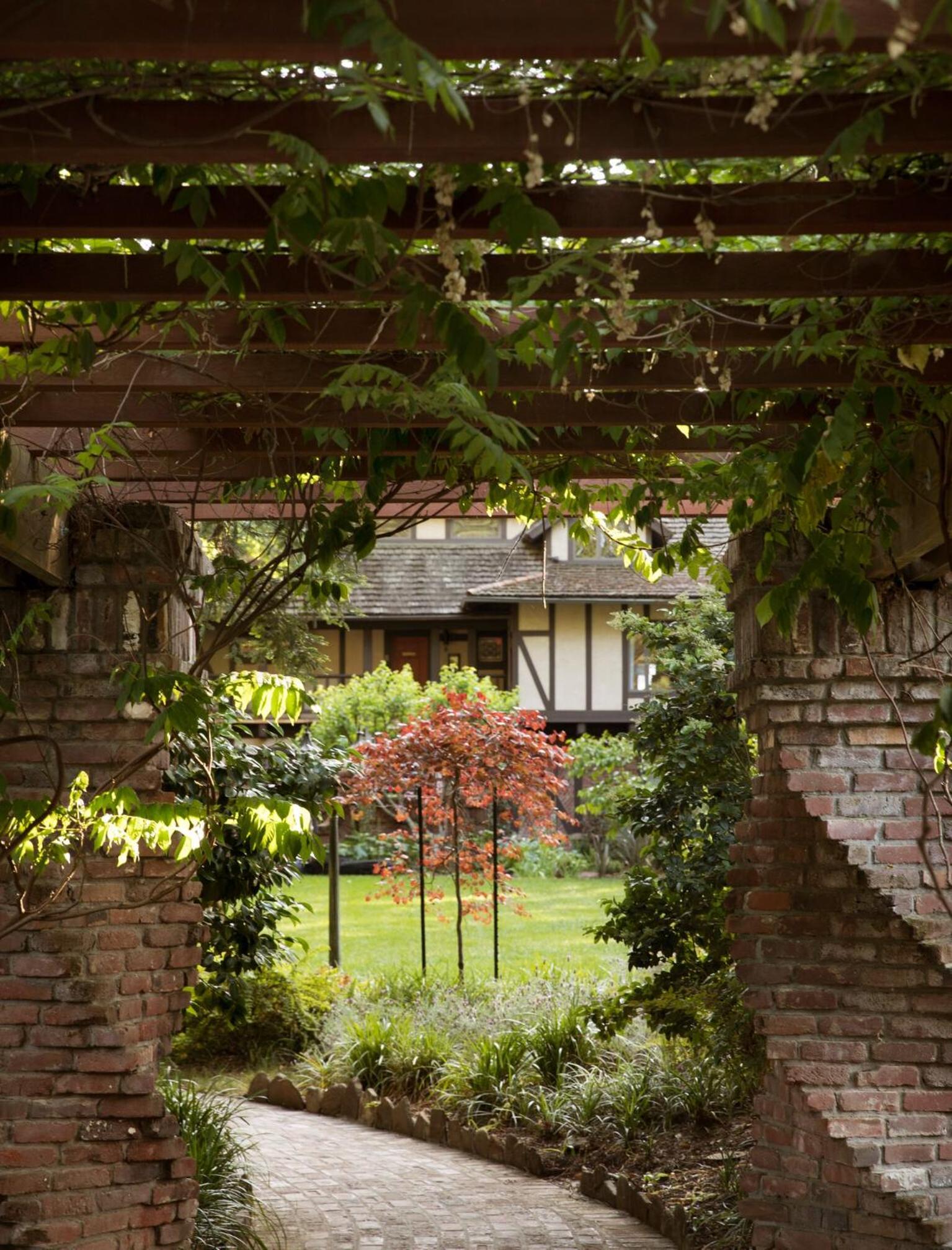 A curved brick walkway seen through a vine-draped wooden pergola leads past foliage toward a Tudor cottage.