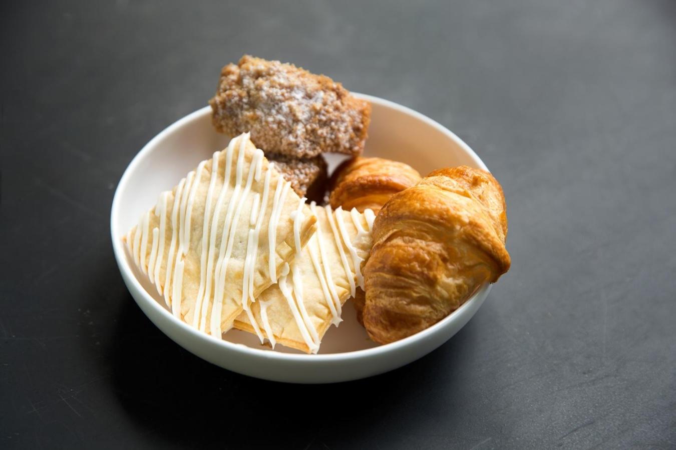 Croissants and iced pastries arranged in a white ceramic bowl on a dark table.