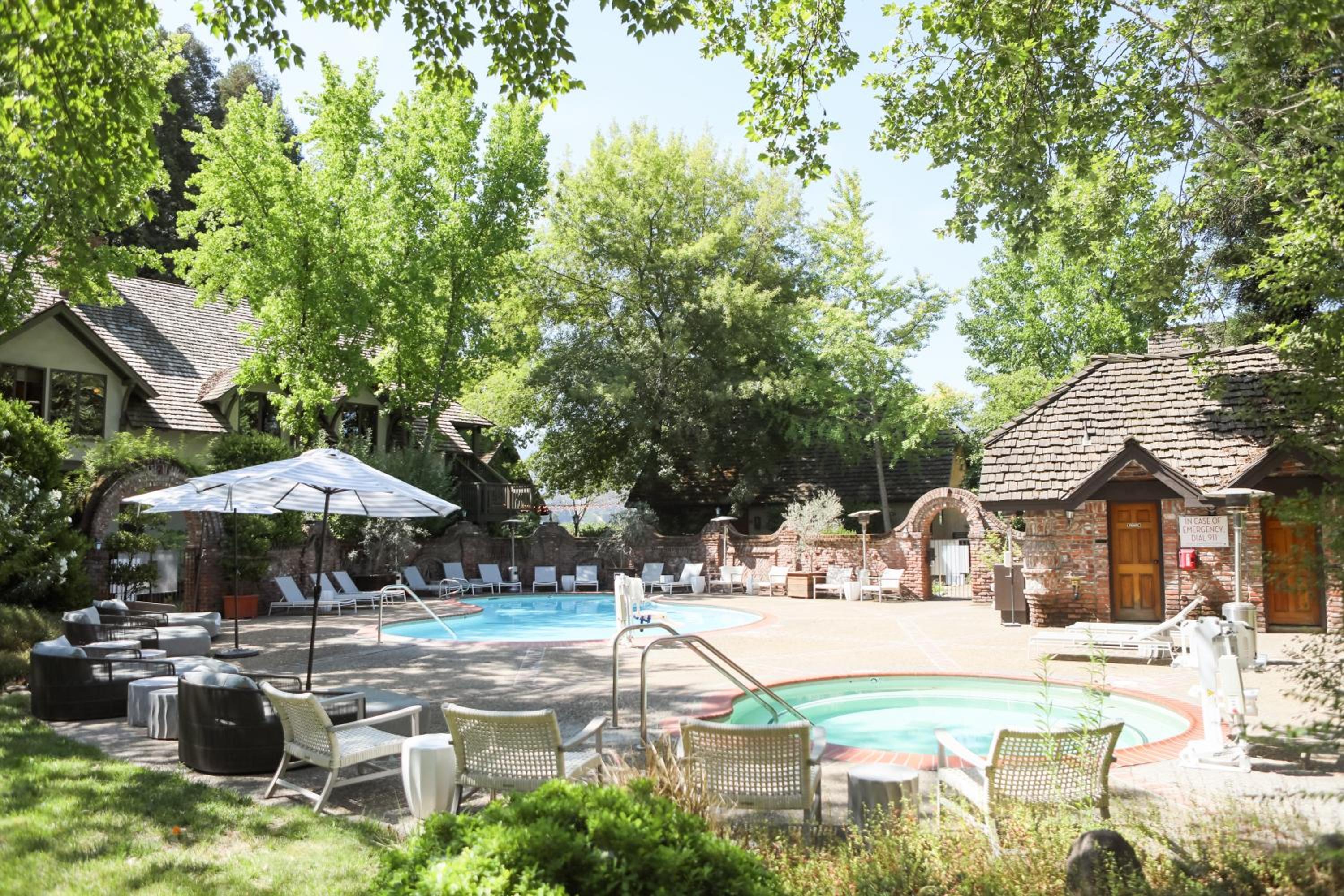 An outdoor swimming pool and circular hot tub bordered by patio lounge seating, brick arches, and tall leafy trees.