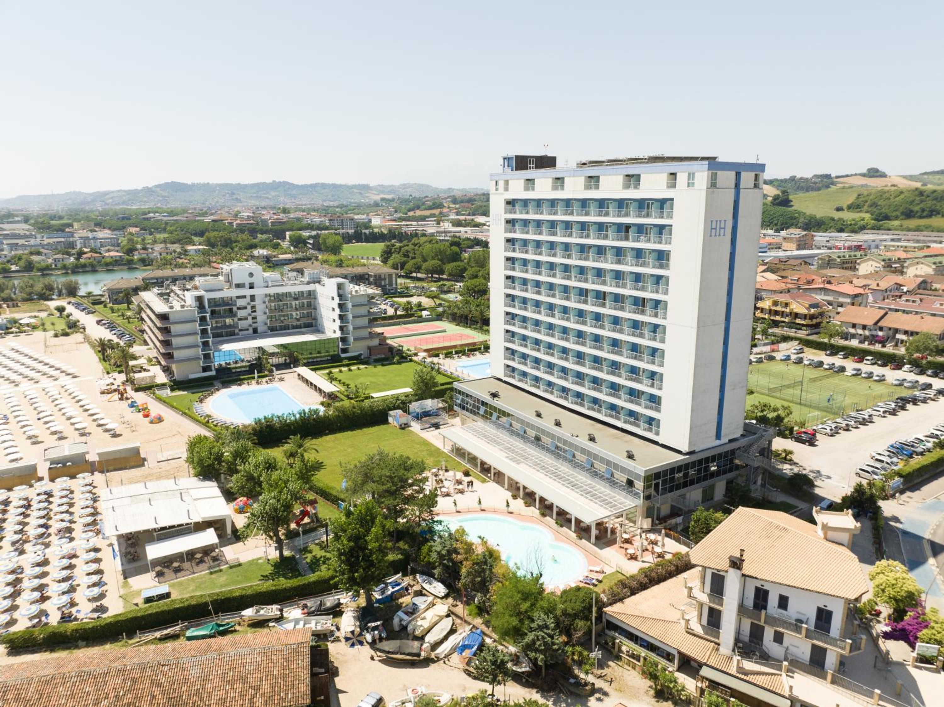 Aerial view of the Hermitage Hotel tower, swimming pools, green lawn, and the coastline.