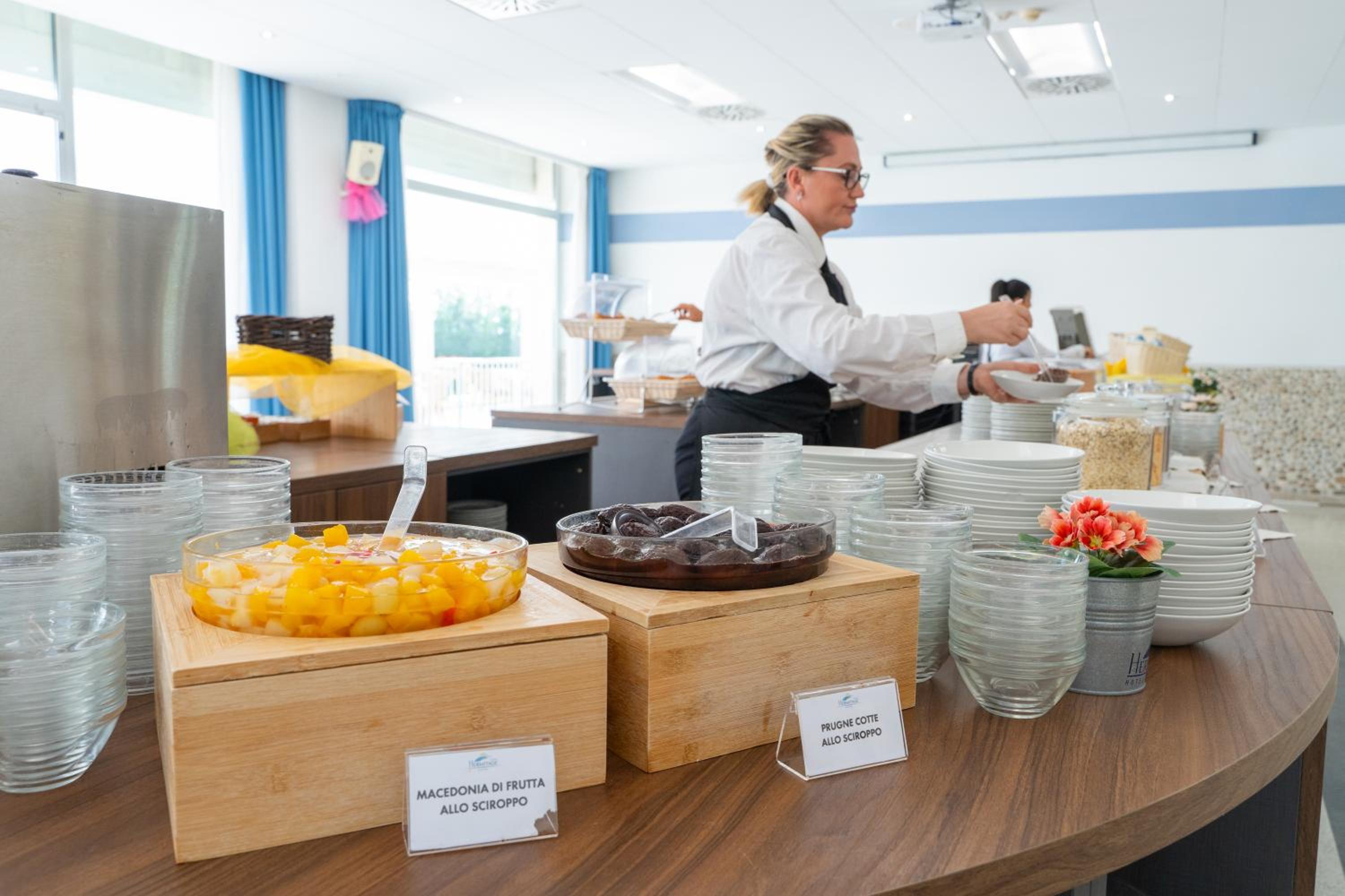 Glass bowls of fruit in syrup sit on wooden stands along a breakfast buffet counter.
