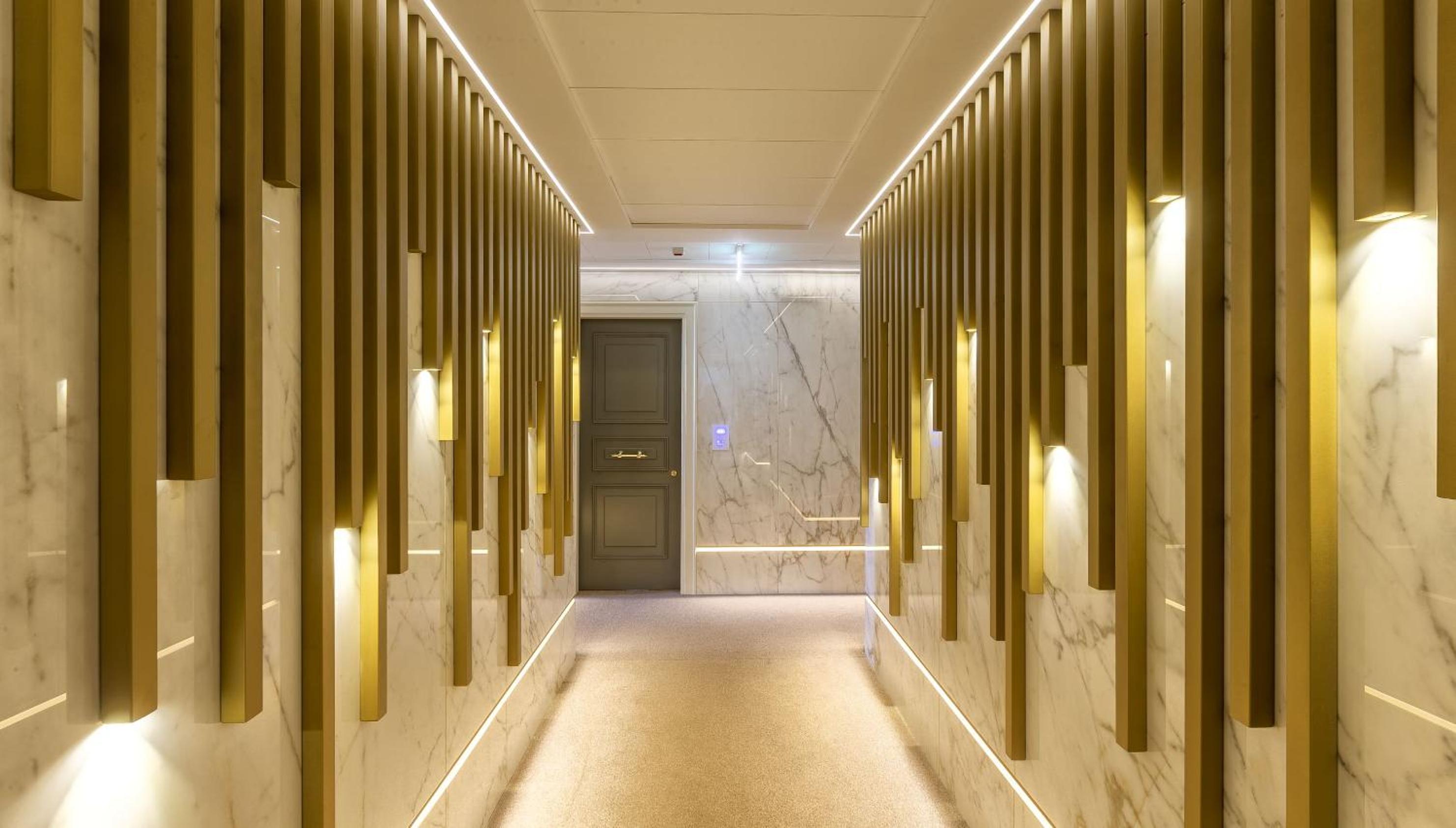 A hallway lined with marble panels, vertical gold slats, and recessed warm lighting leading to a dark door.