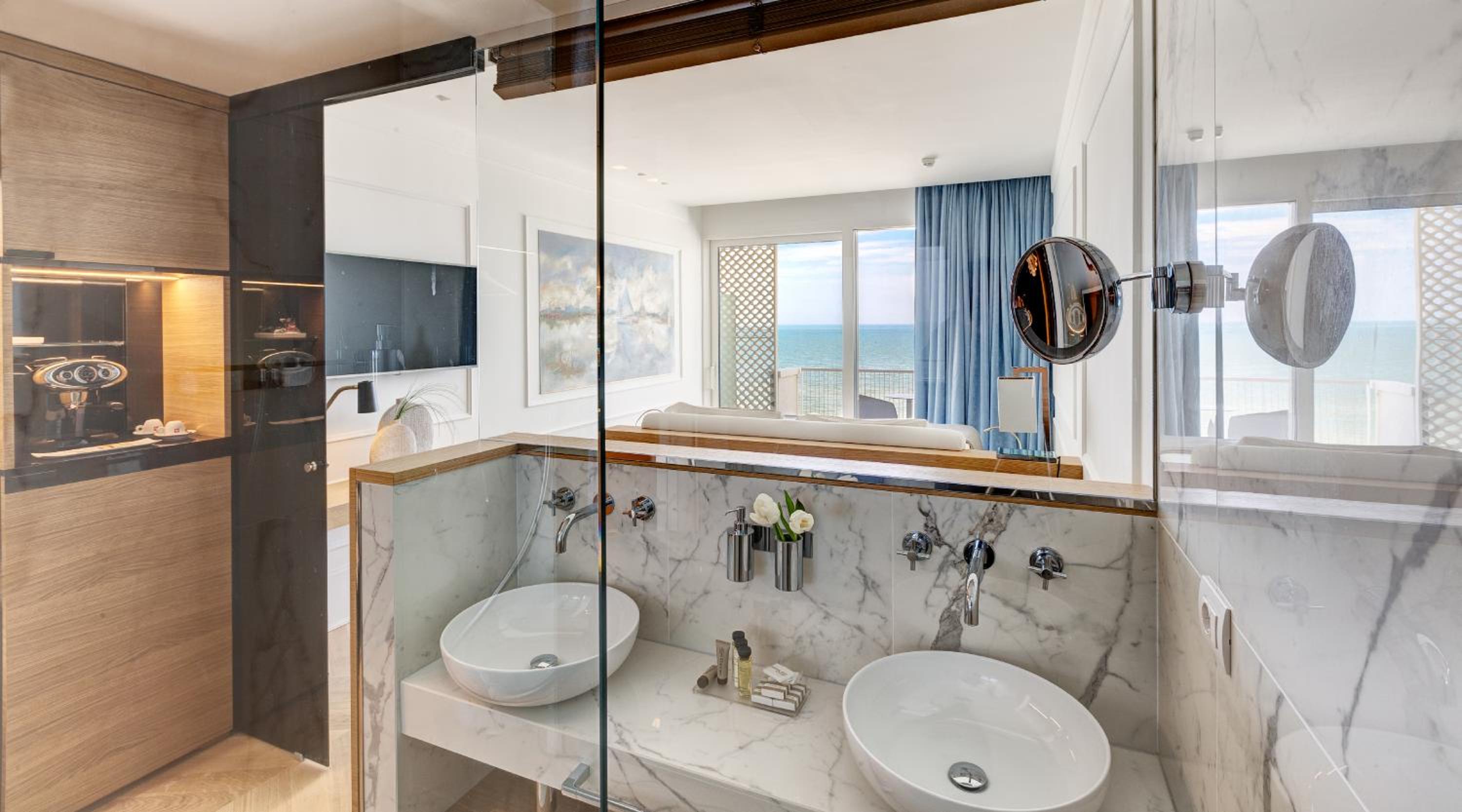 Marble bathroom vanity with round vessel sinks, reflecting a bright bedroom and the sea through large windows.