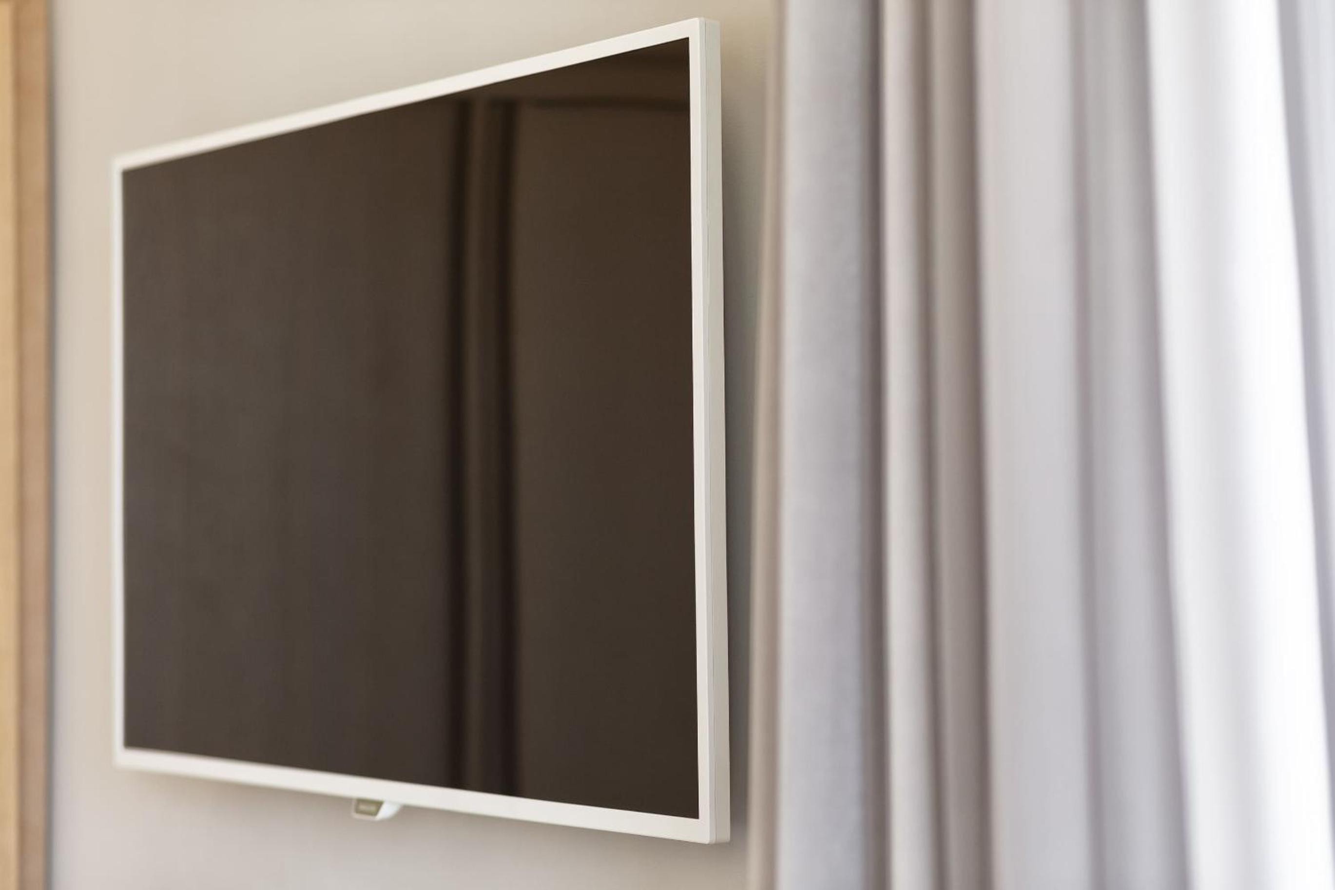 A flat-screen television mounted on a neutral wall beside gathered sheer curtains.