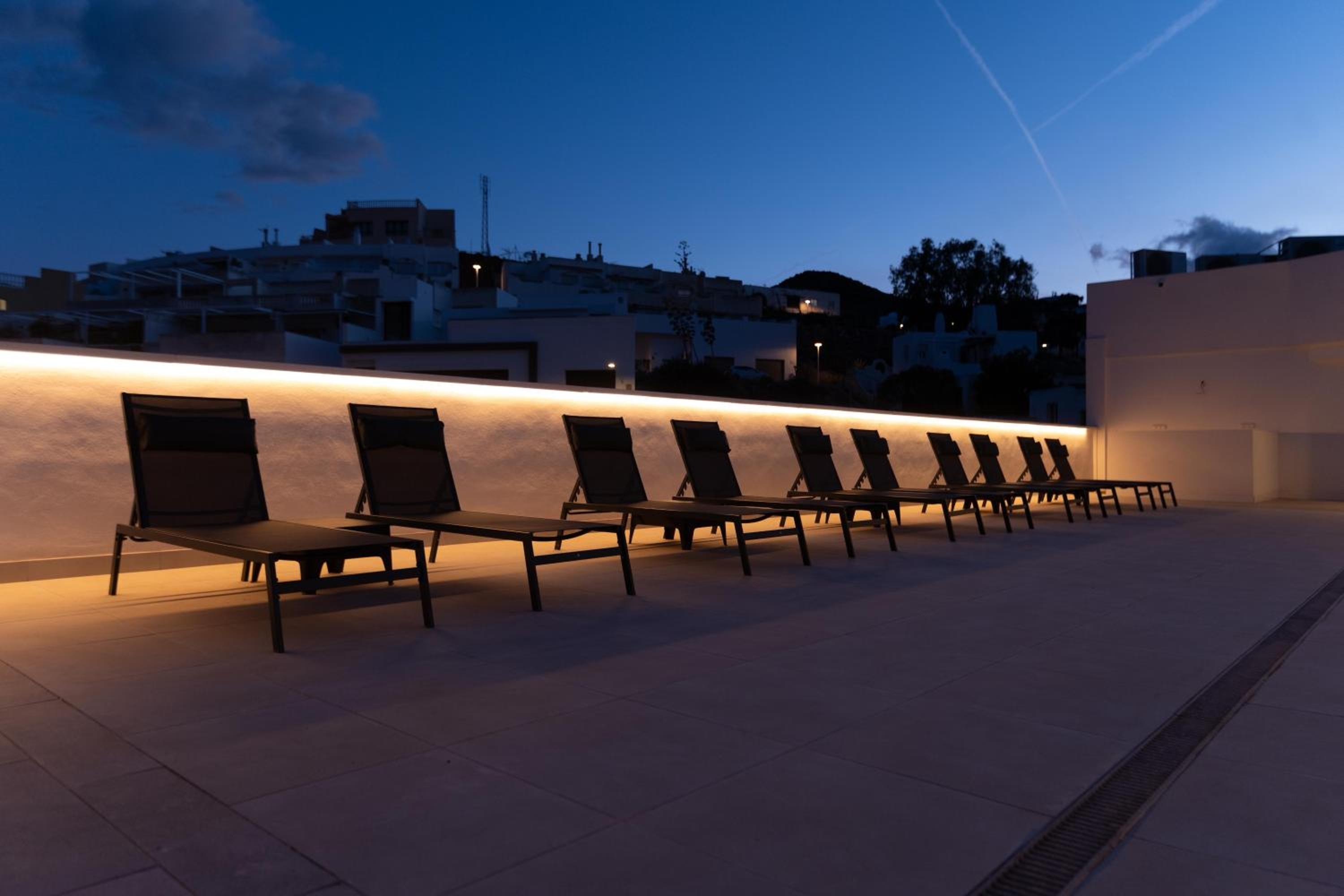 Sun loungers set beside an illuminated low wall on a paved rooftop terrace at twilight.