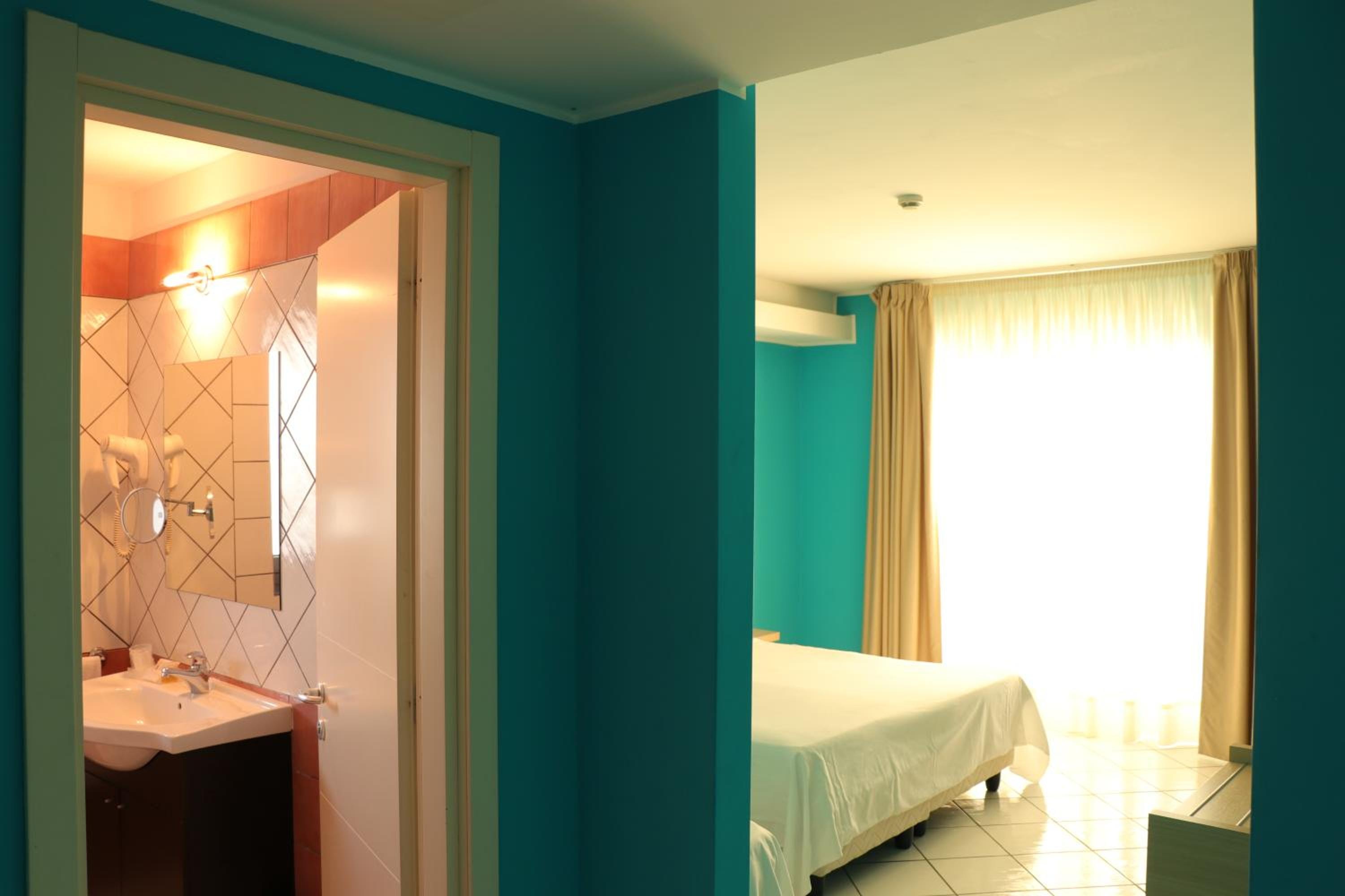 A bright bedroom with turquoise walls and a white-sheeted bed, viewed beside an open doorway to a tiled bathroom.