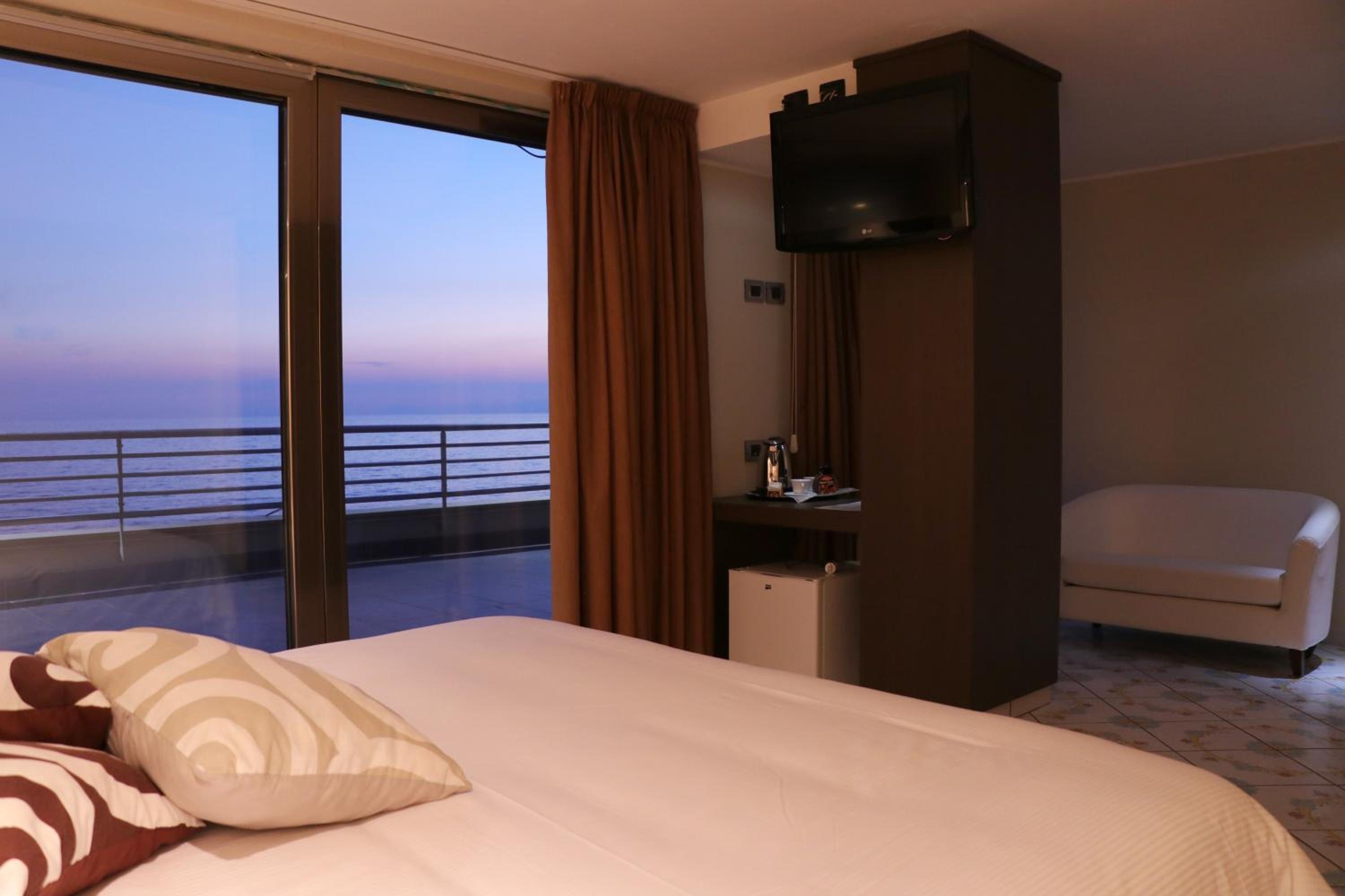 A bedroom with a bed in the foreground and sliding glass doors opening to a balcony with views of the sea at dusk.