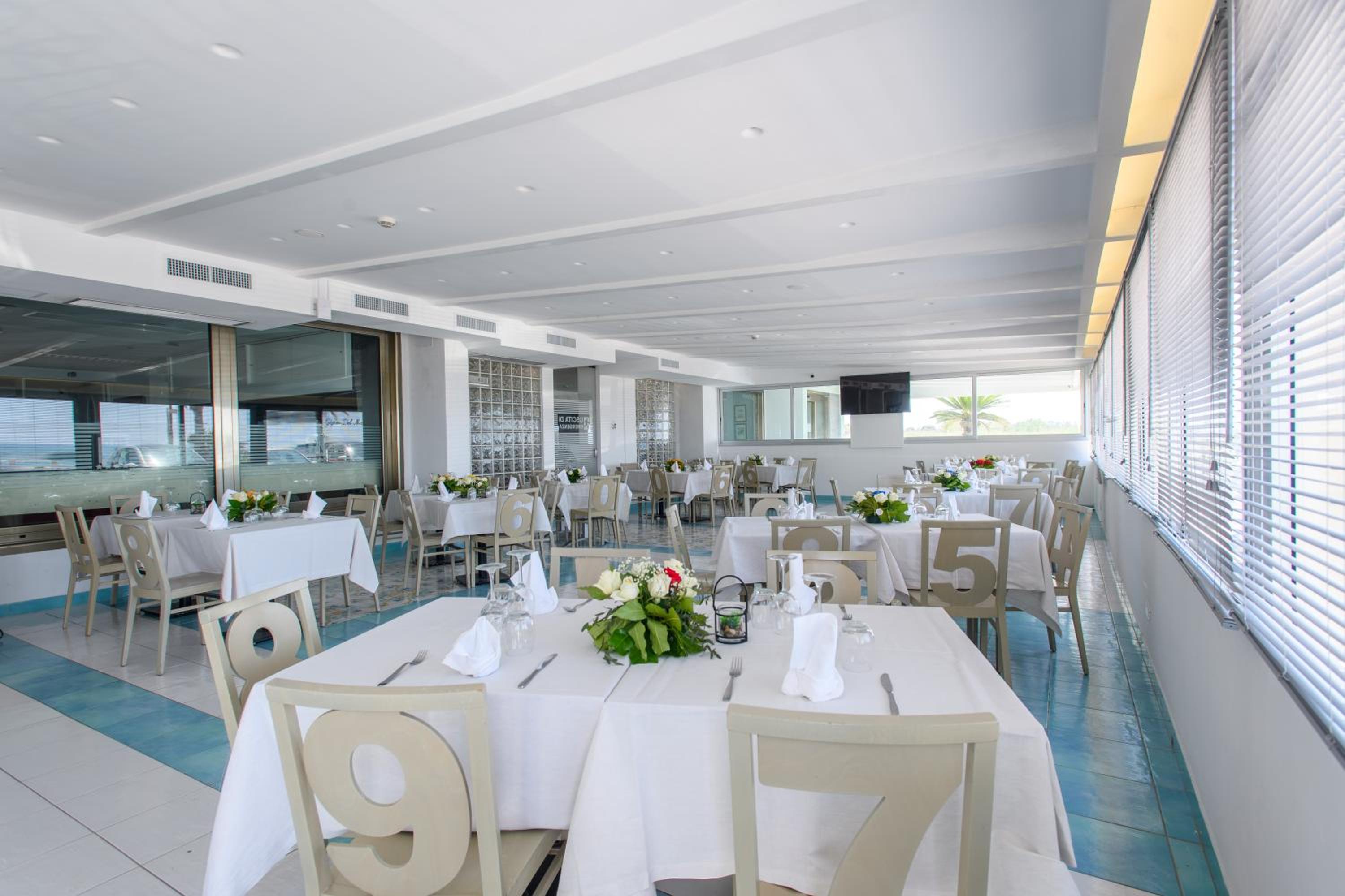 A brightly lit restaurant dining hall with white tables, patterned tile flooring, and windows fitted with blinds.