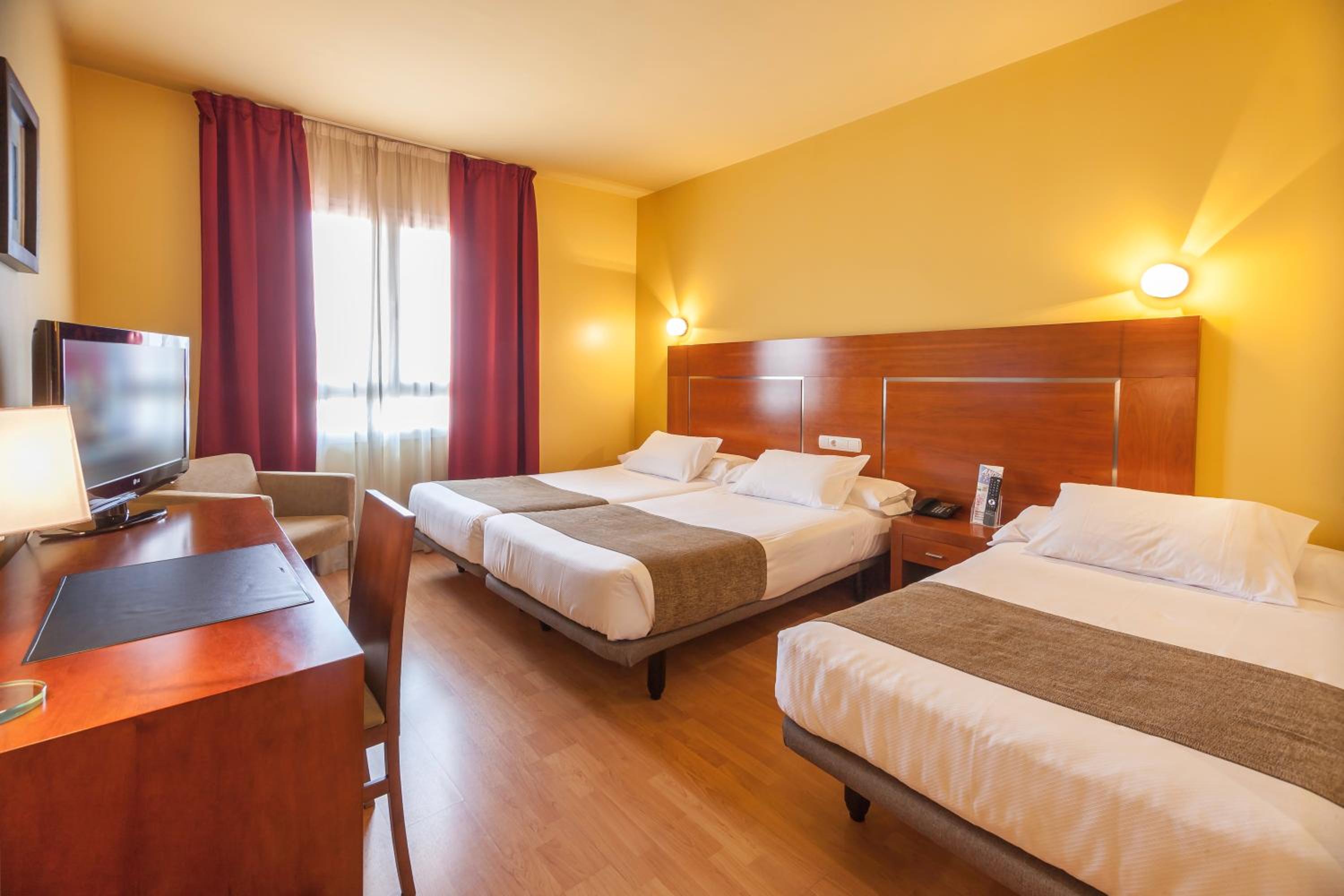 A bedroom with yellow walls, beds along a wood headboard, a work desk, and red curtains.