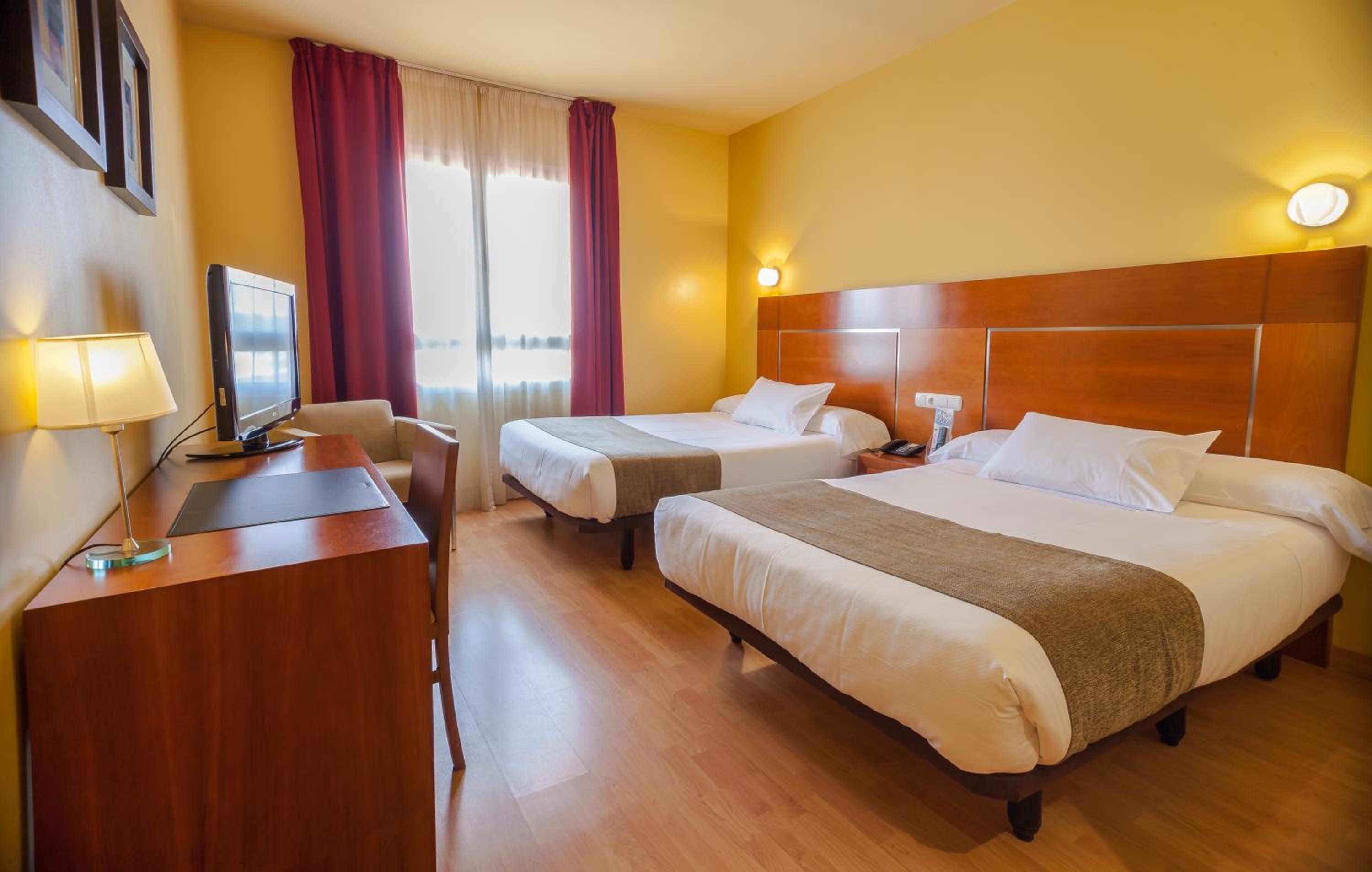 A hotel room with beds, yellow walls, a wood desk with a television, and red window drapes.