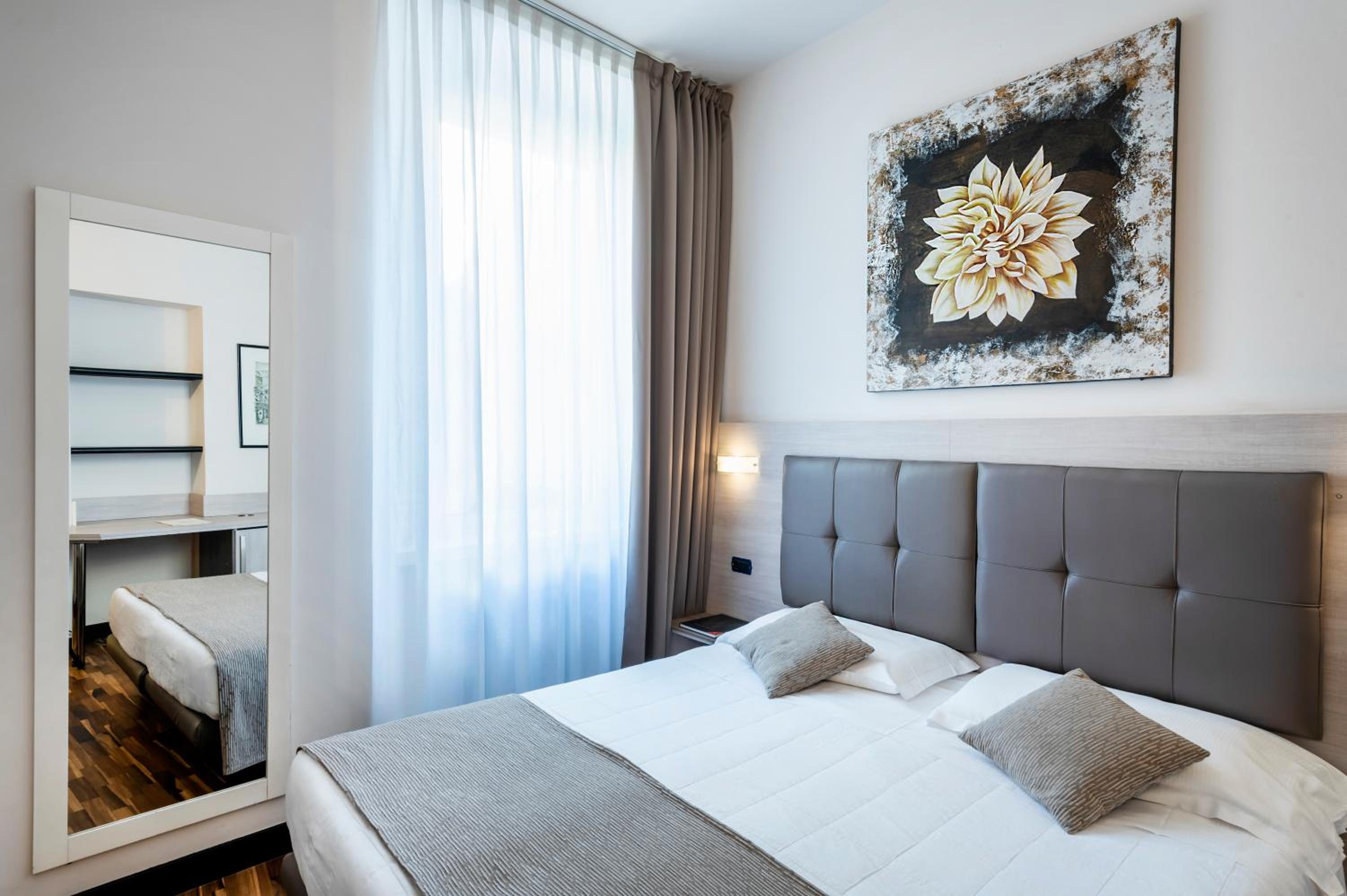 Bedroom with a padded leather headboard, floral painting, sheer curtains, and full-length mirror.