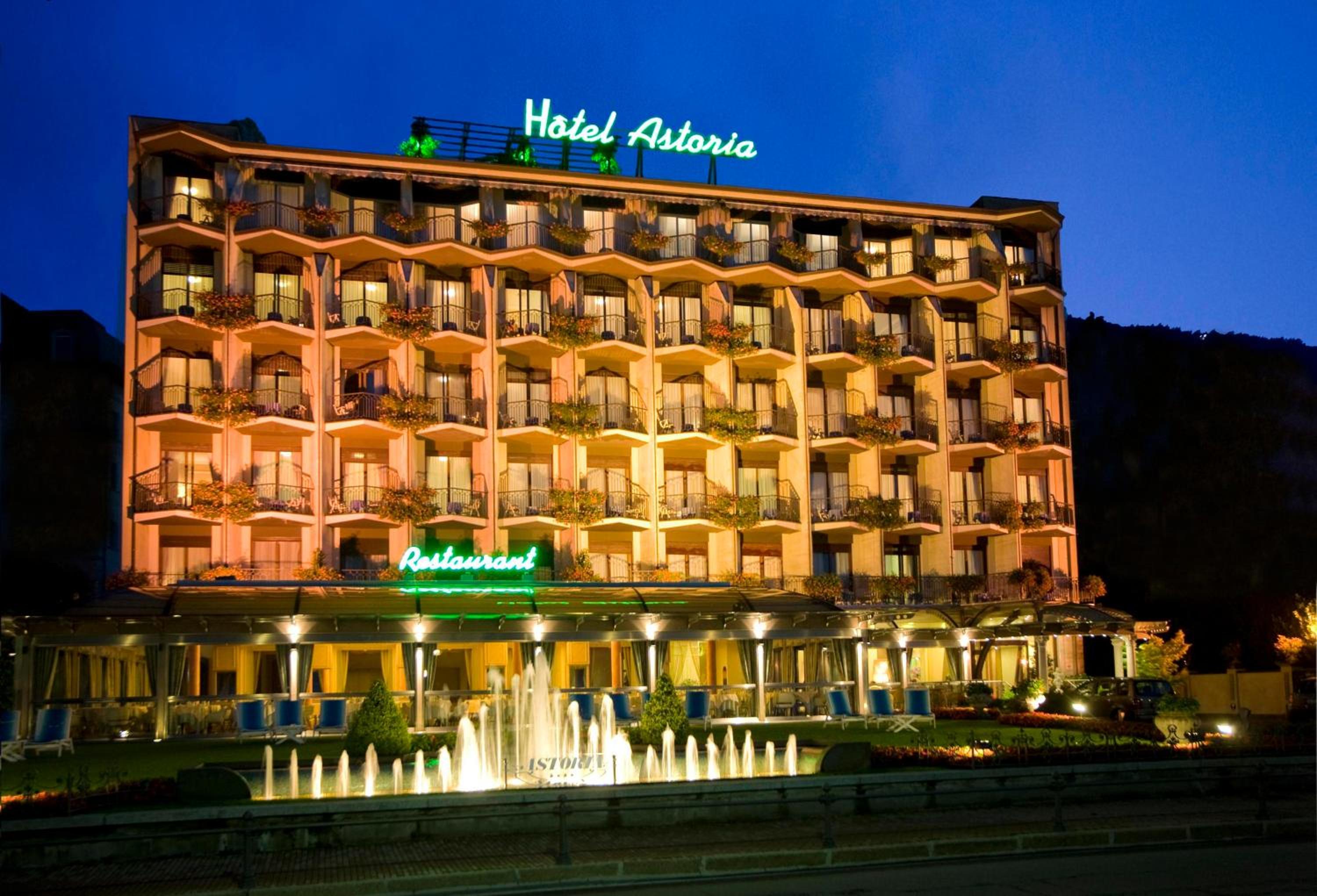 Illuminated multistory facade of Hotel Astoria at dusk, with balcony boxes and a water fountain in front.