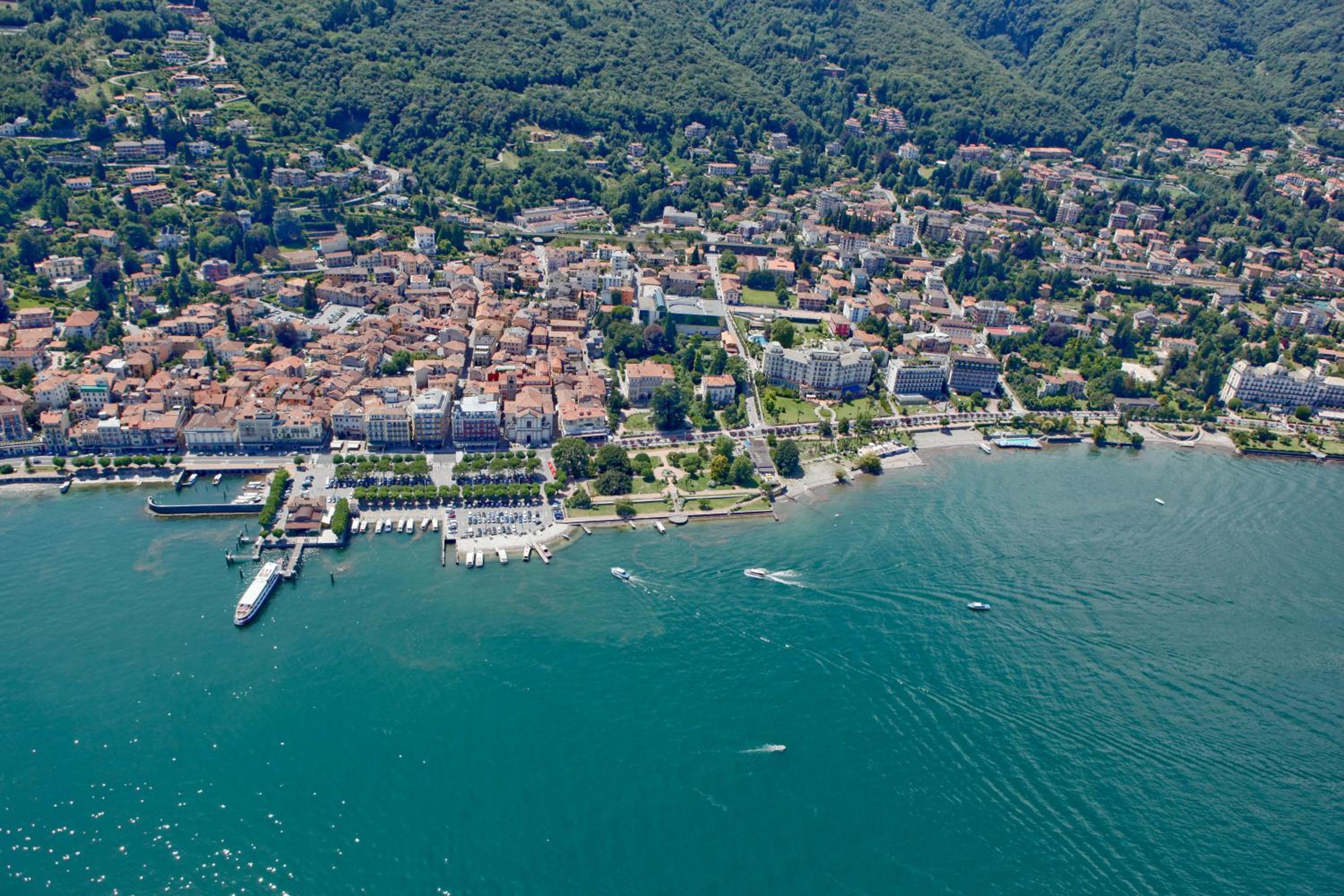 Aerial view of a lakeside town along turquoise water with boats, docks, and forested mountain slopes behind.