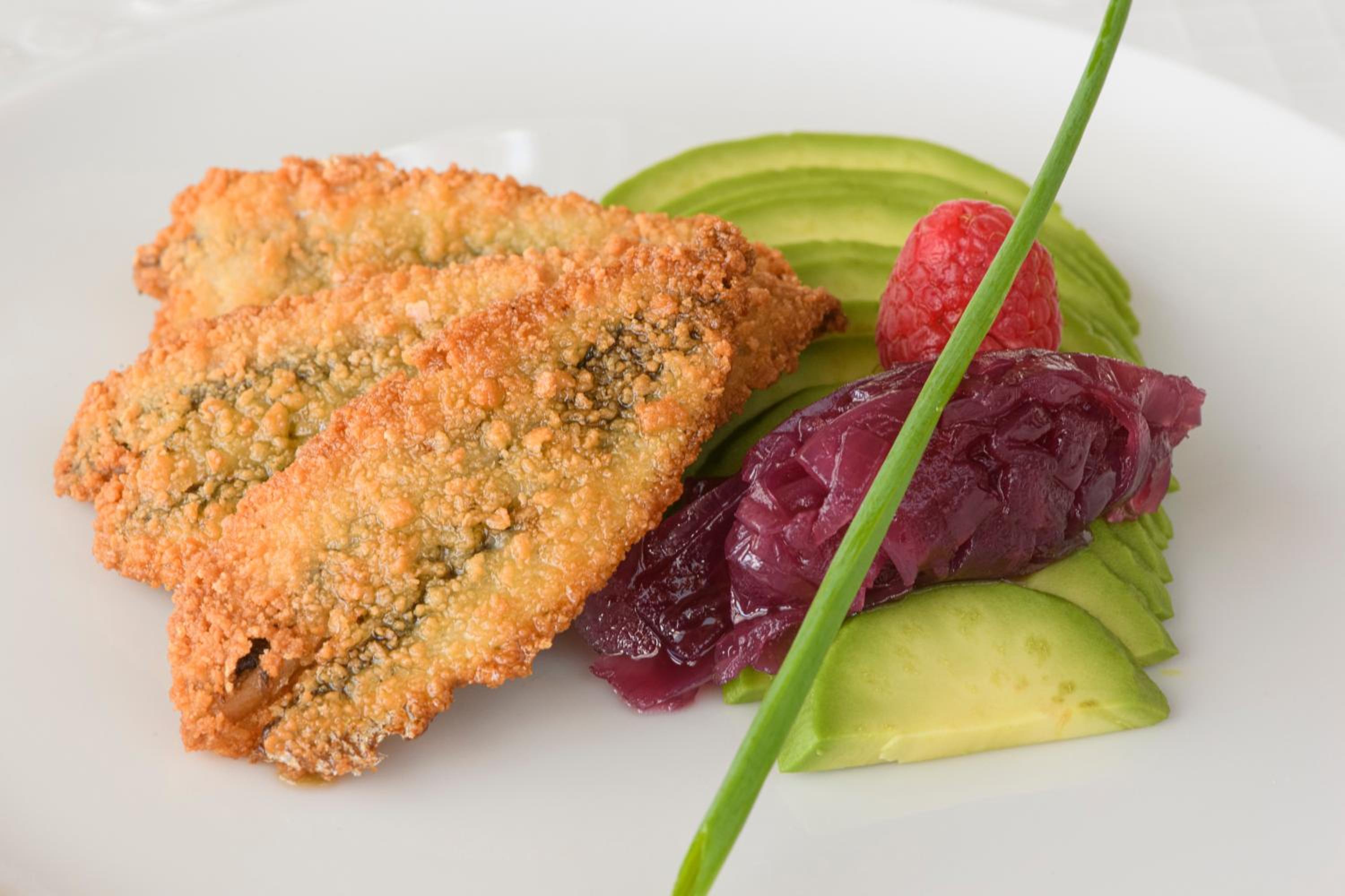 Plated breaded fish fillets served with sliced avocado, red onion compote, a raspberry, and a chive garnish.