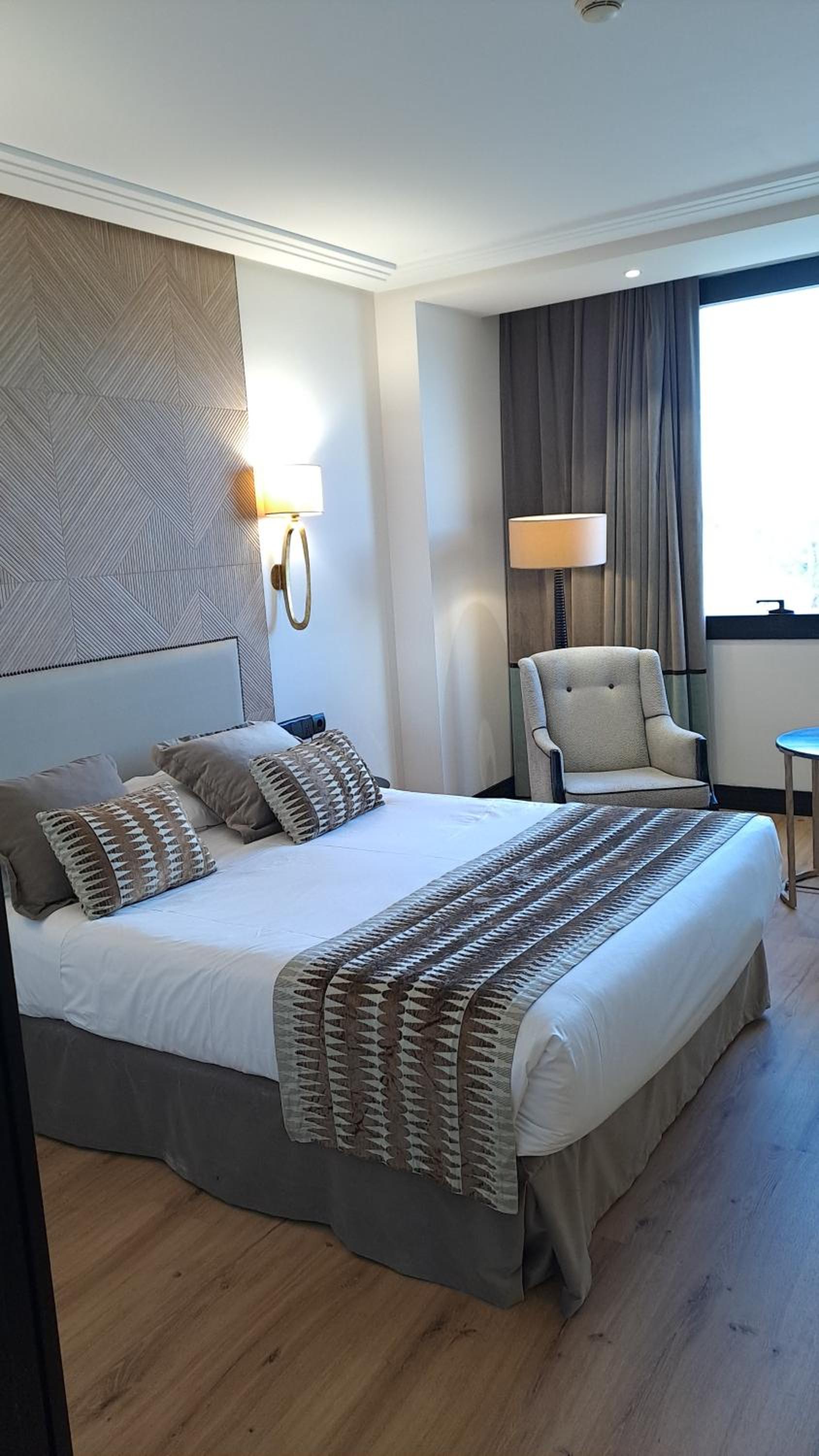 A made bed with patterned accent pillows and runner, beside an upholstered armchair and draped window.