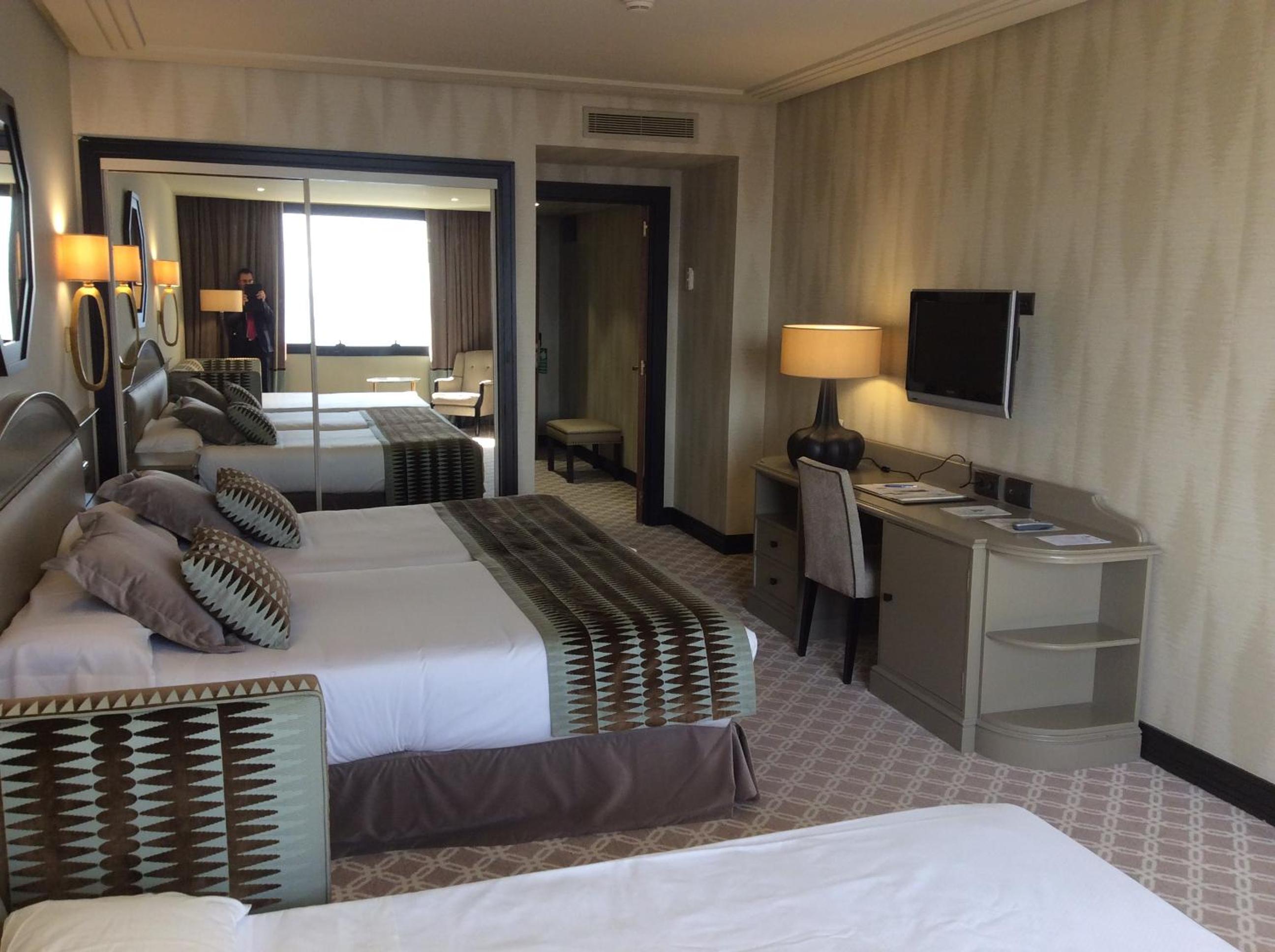 A hotel room with a work desk, wall-mounted television, and mirrored wardrobe reflecting the interior.