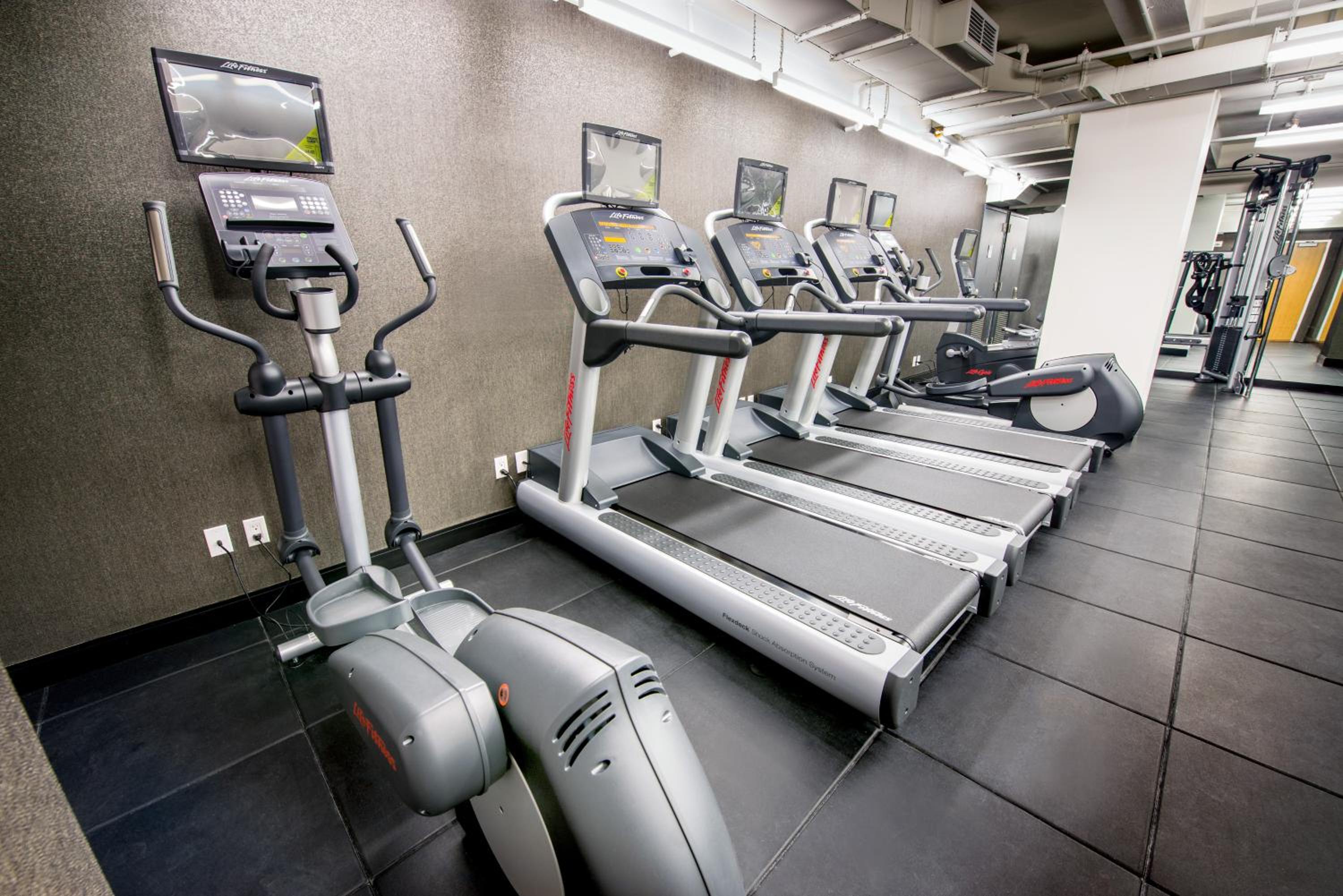 A gym with treadmills and elliptical equipment arranged along a textured gray wall.