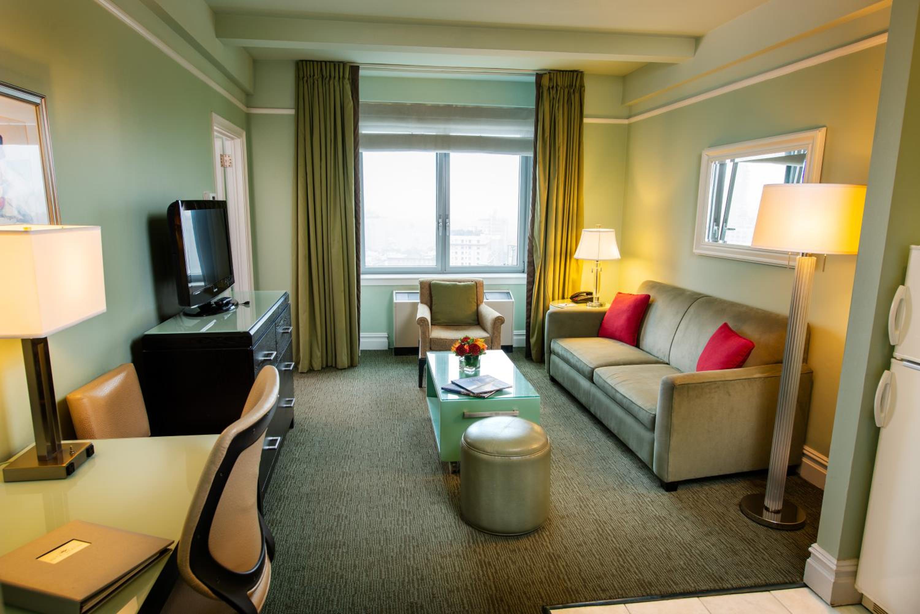 A suite living room with pale green walls, carpet, a sofa with red accent pillows, and a window.