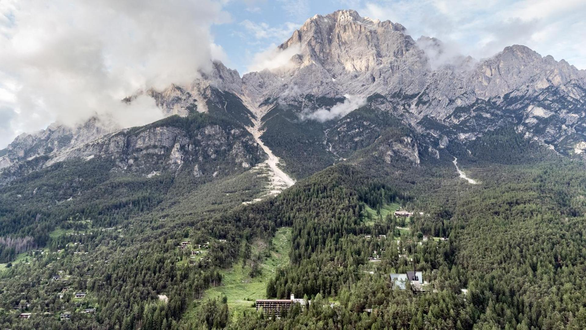 Dense pine forests blanket the mountain slopes beneath a rocky alpine peak shrouded in passing clouds.