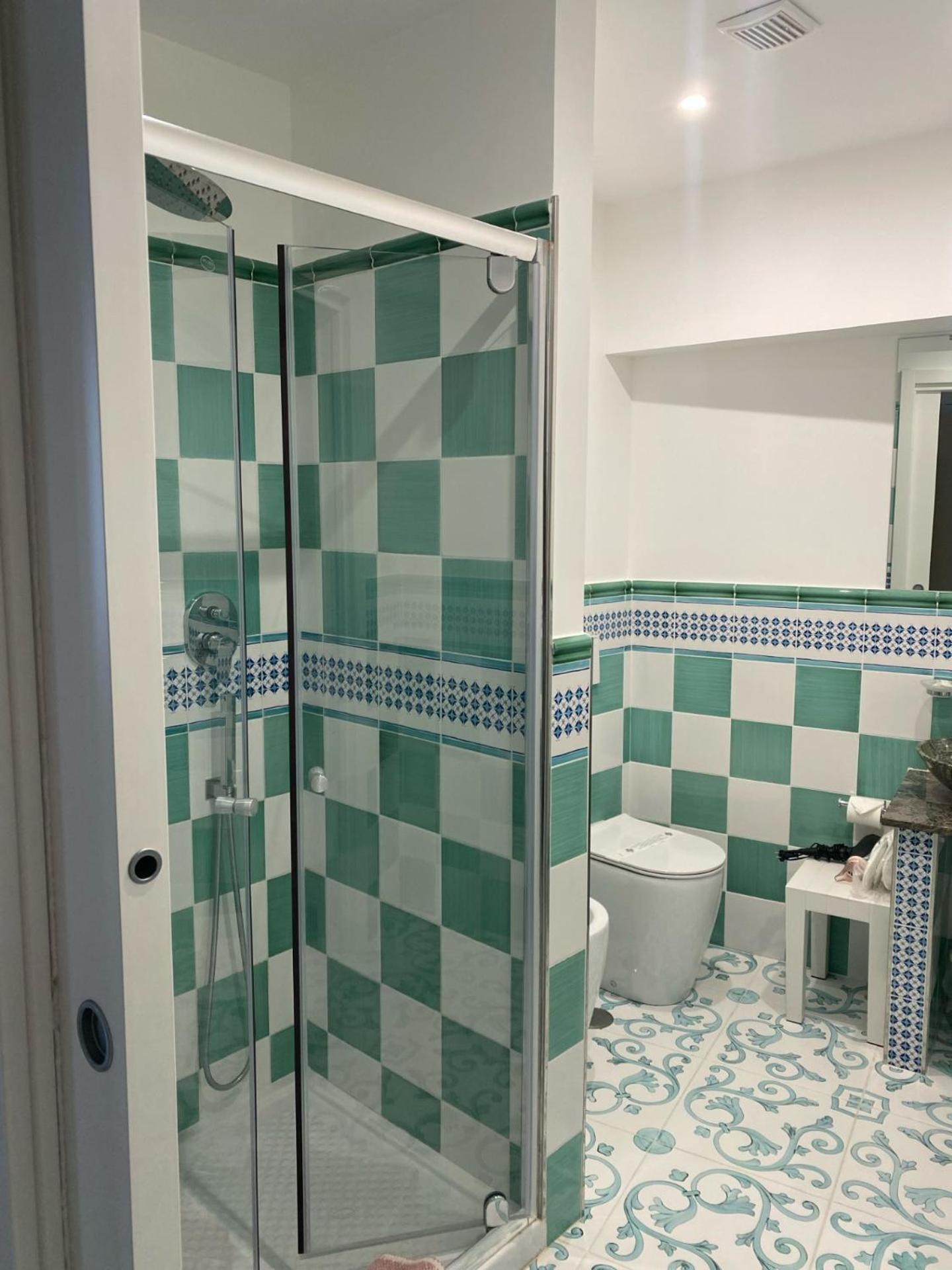 A bathroom with green checkered wall tiles, patterned floor tiles, a glass shower stall, and a white toilet.