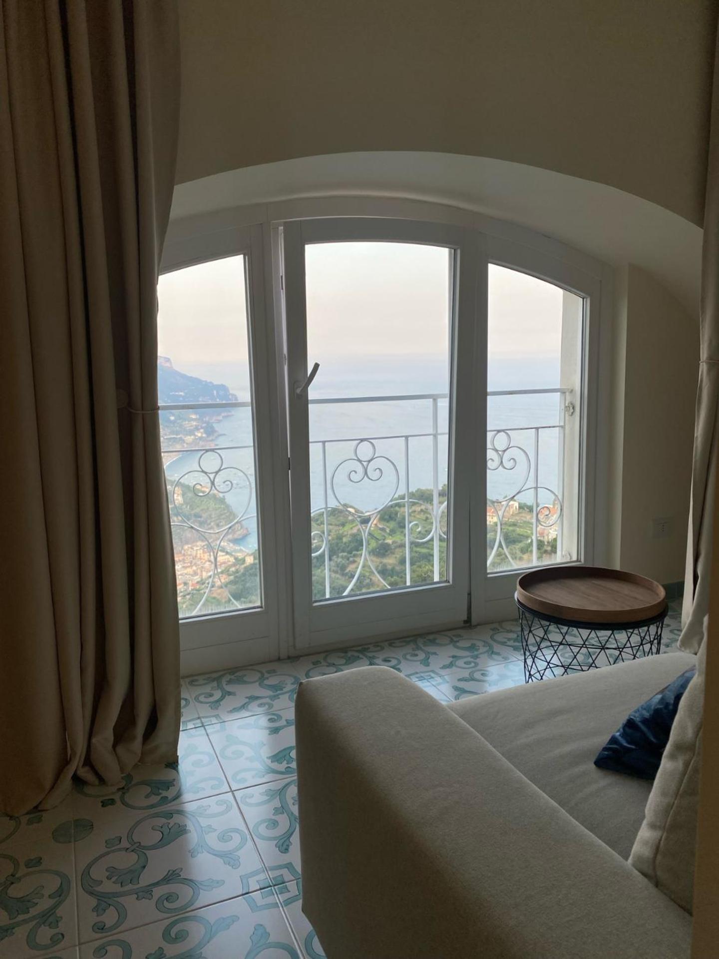 An arched window with white railings framing a view of the sea and coastline beside a sofa and patterned floor.