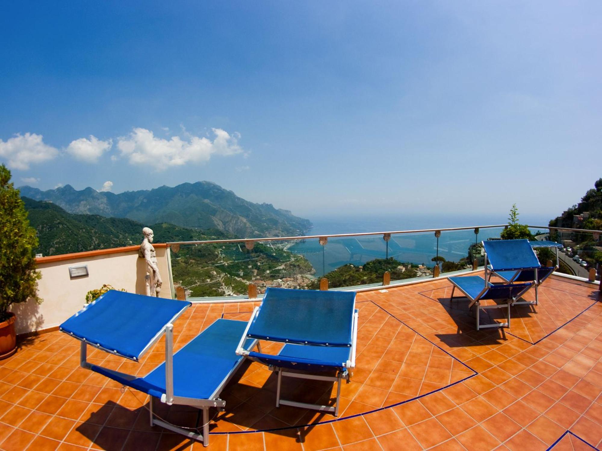 A terracotta tiled terrace with blue sun loungers and a glass railing overlooking mountains and the sea.