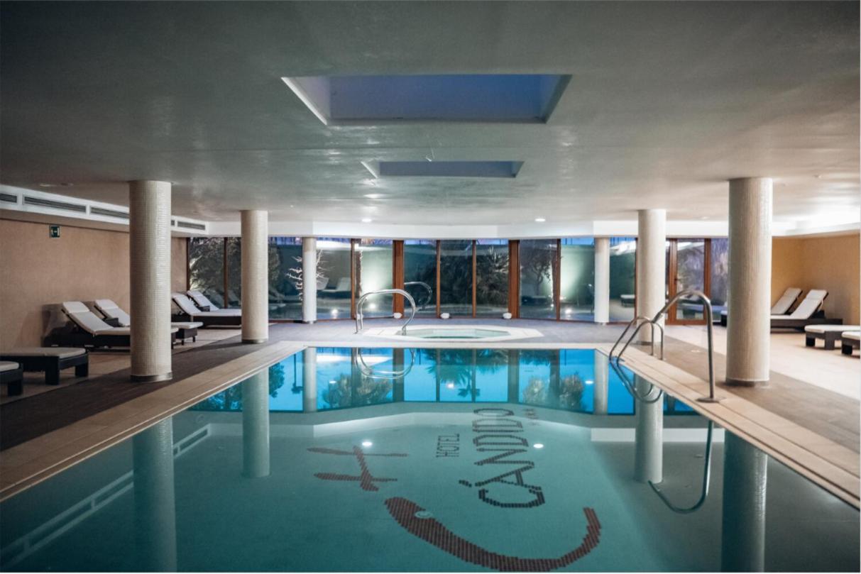 An indoor swimming pool at Hotel Cándido with tiled columns, sun loungers, and glass doors looking outdoors.