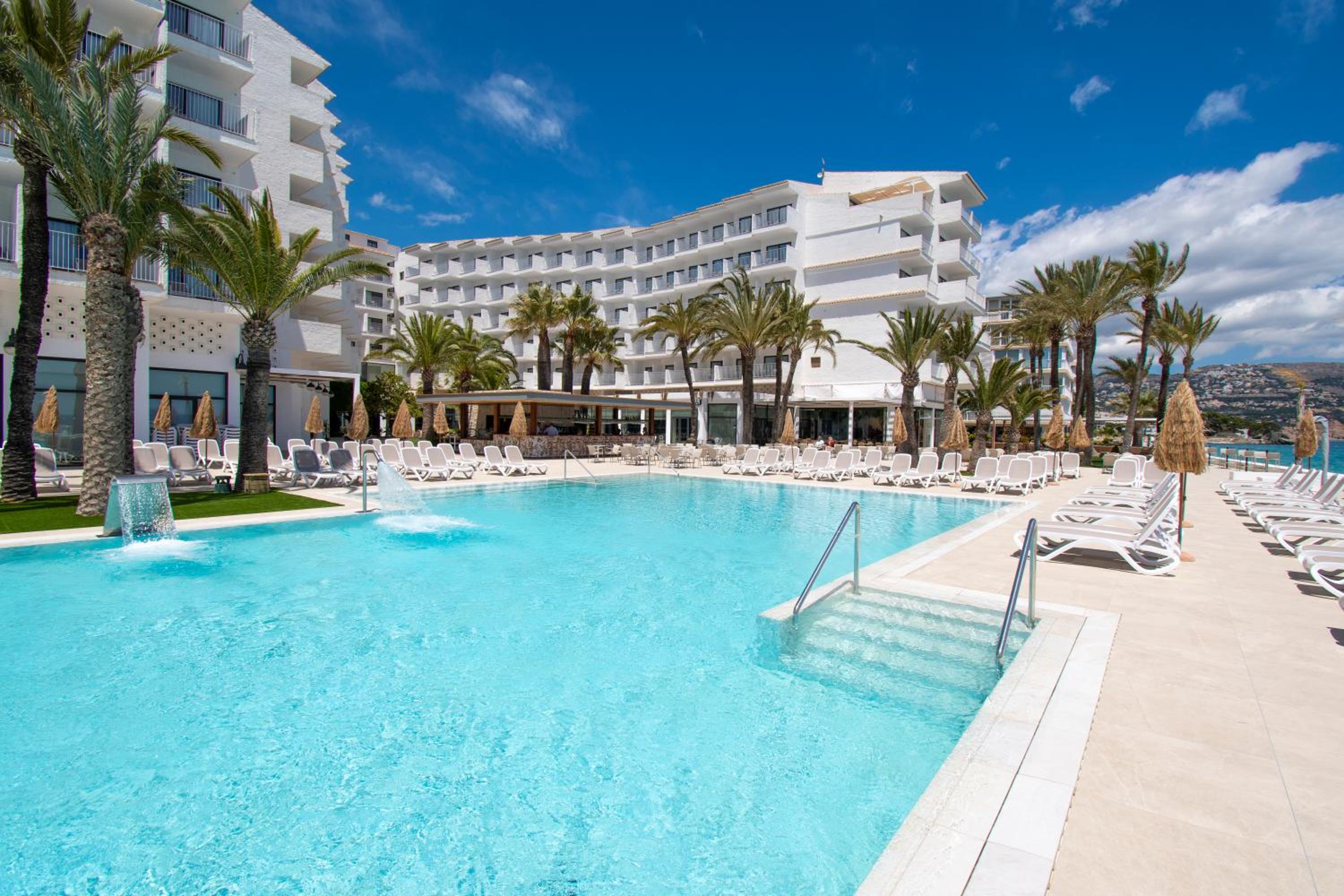 An outdoor swimming pool at Hotel Cap Negret, lined with white sun loungers and palm trees under a sunny sky.