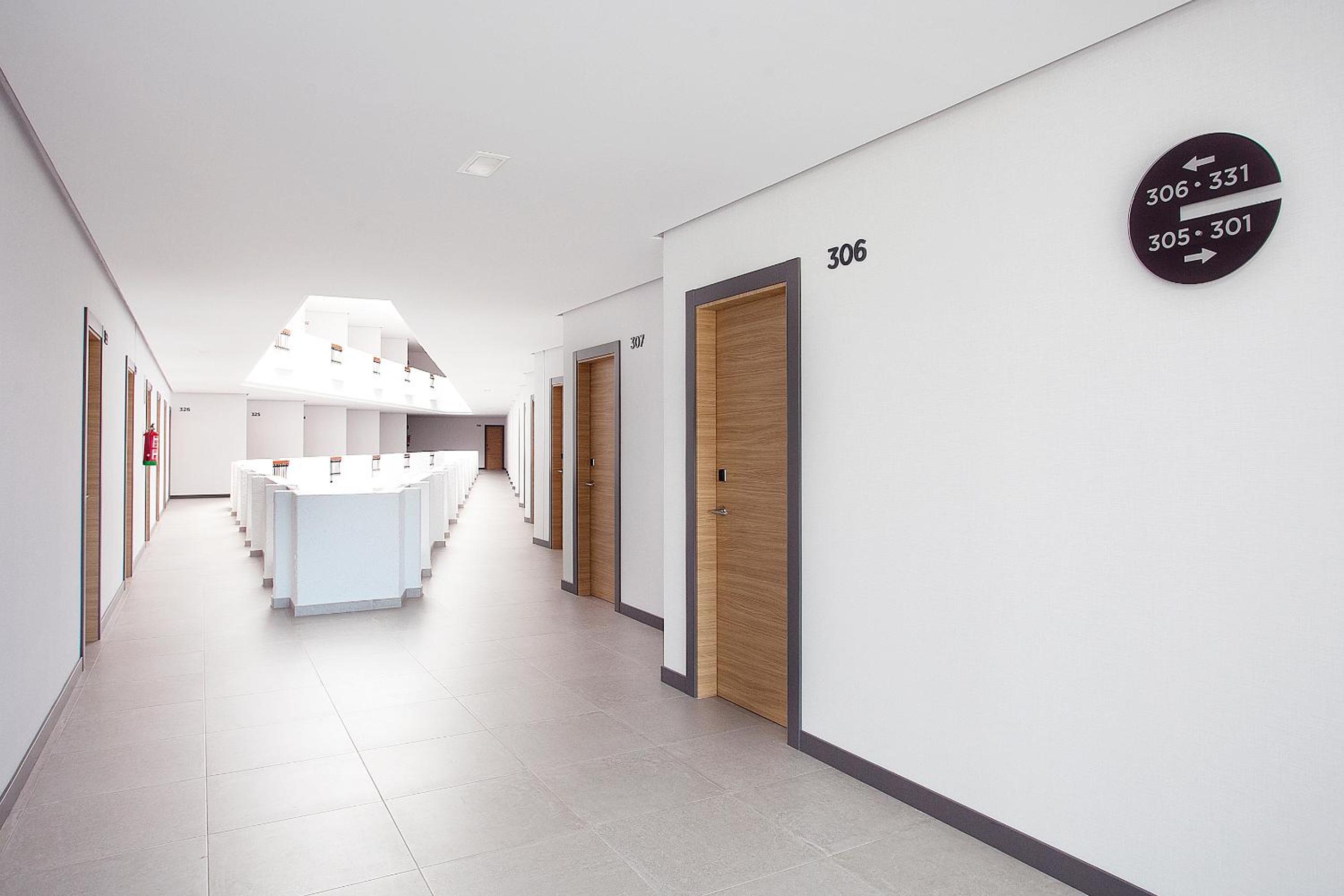 A bright hotel hallway with white walls, tiled floors, wooden doors, and directional signage.