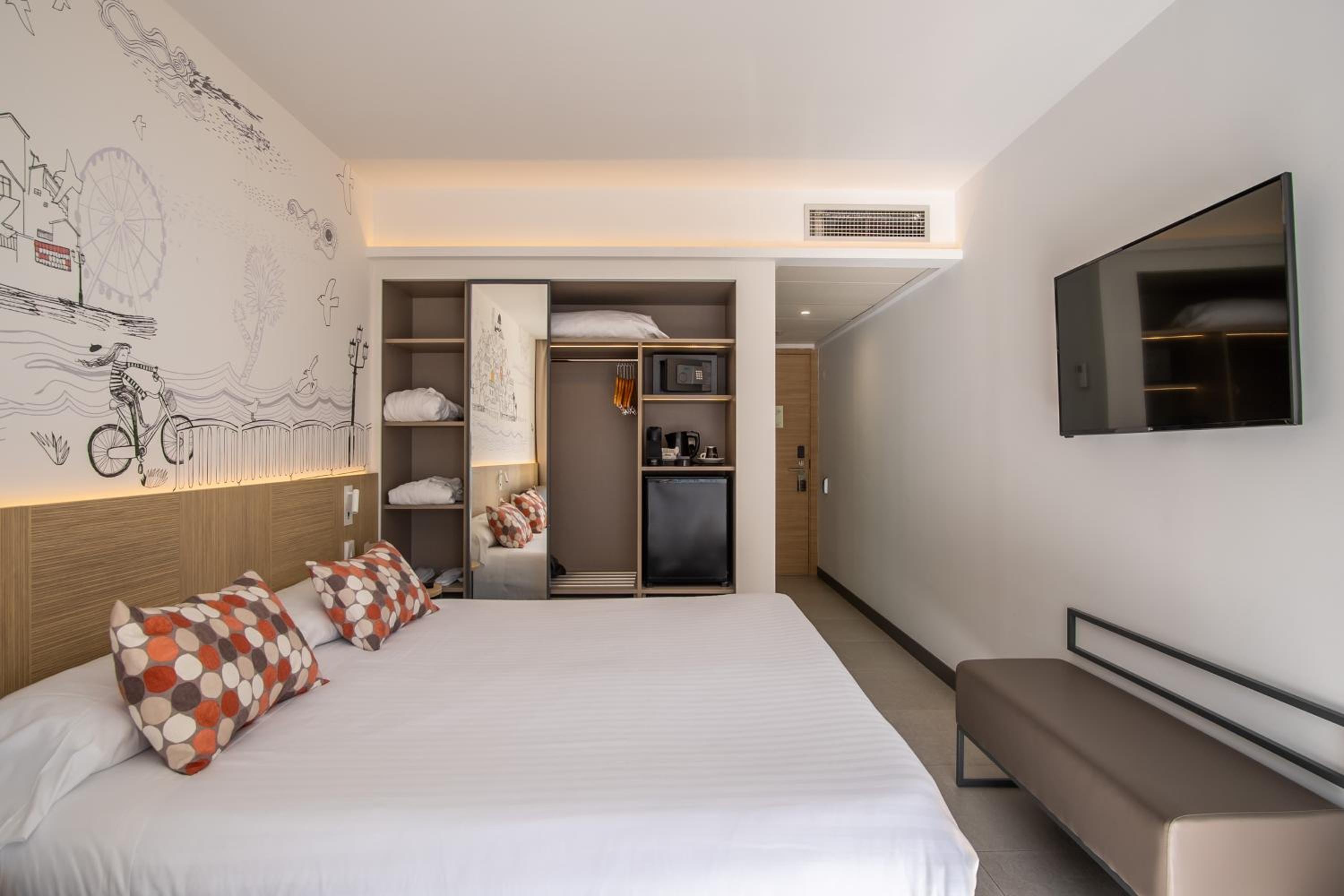 A hotel bedroom with a large bed, patterned pillows, an illustrated mural, and a mounted television.