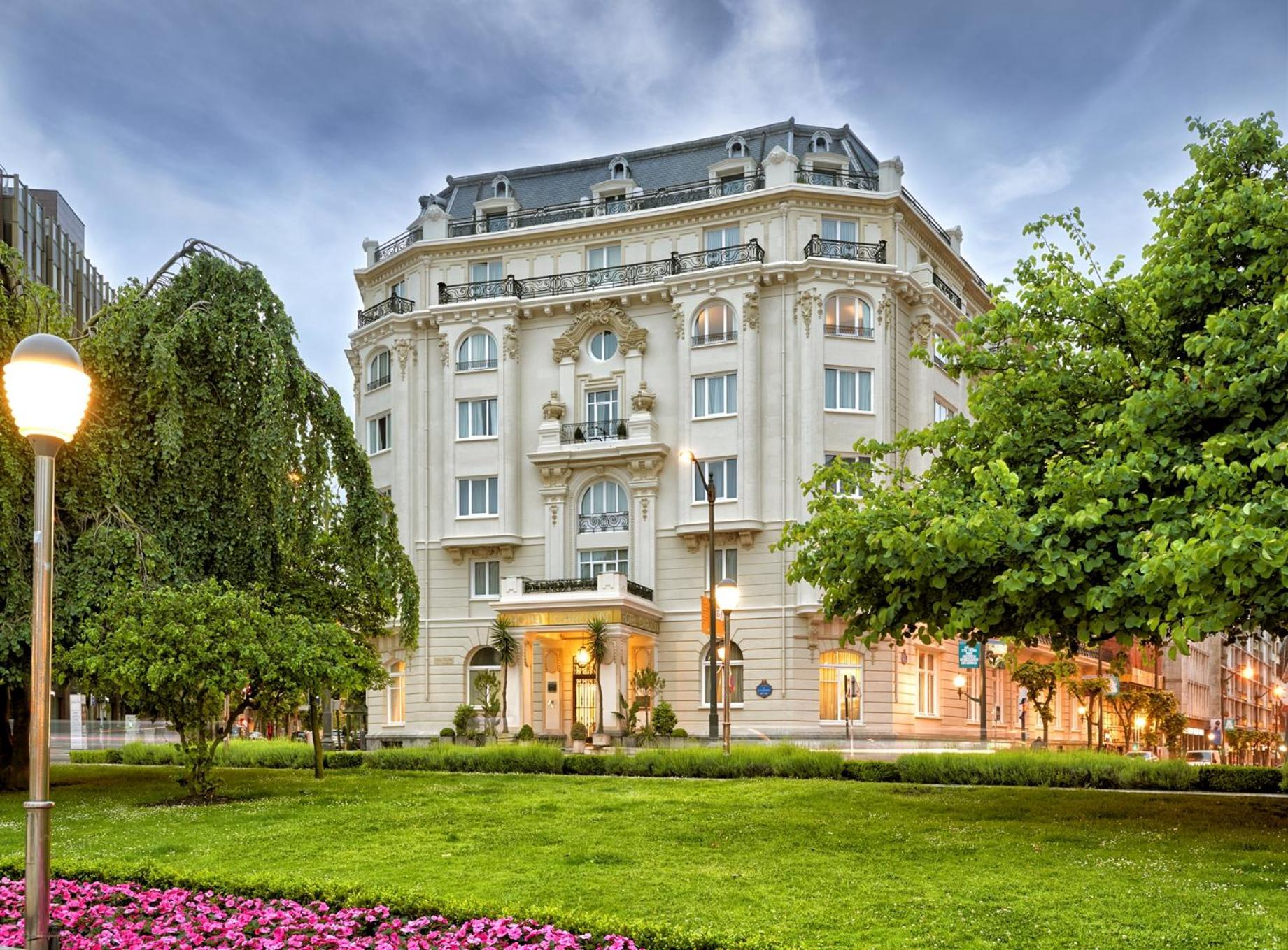 The stone facade of Hotel Carlton stands behind a manicured green lawn, flowering plants, and leafy trees.